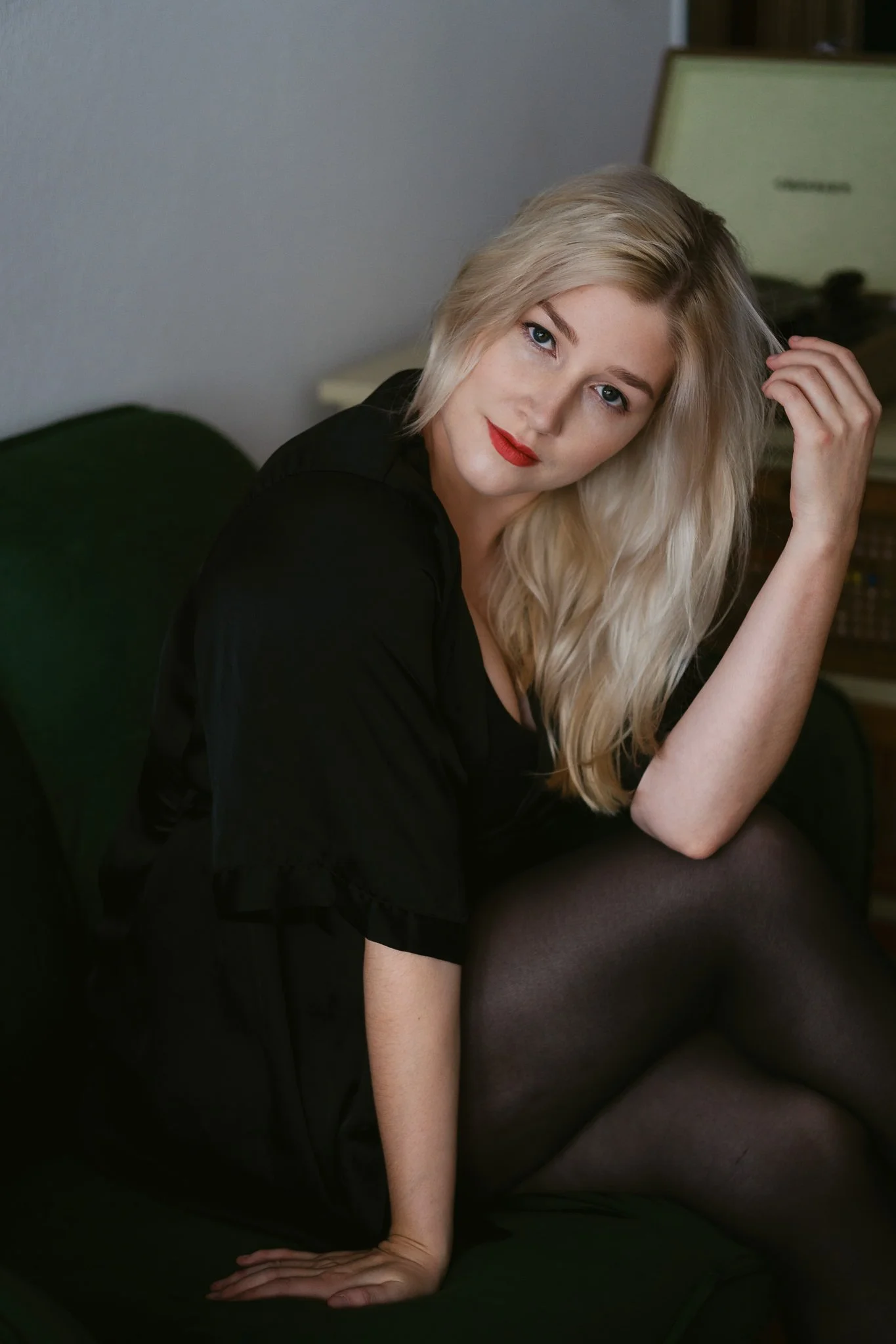 Junge blonde Frau sitzt auf einem grünen Sofa, trägt schwarze Kleidung und roten Lippenstift, schaut in die Kamera, streicht sich durch die Haare.