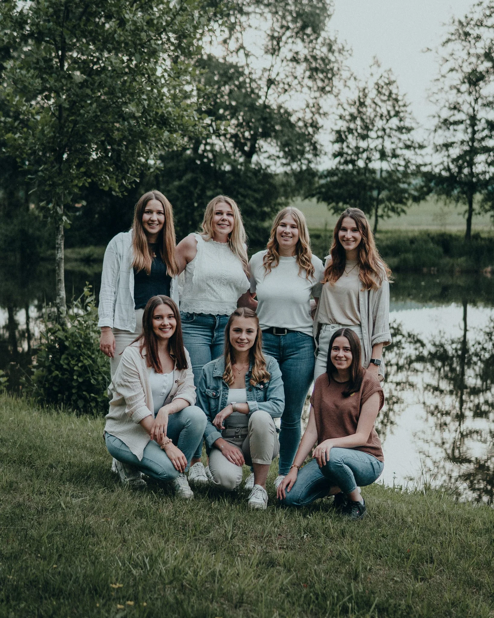 Gruppenfoto von acht jungen Frauen in casual Kleidung, am Ufer eines Sees, umgeben von Bäumen, an einem bewölkten Tag.