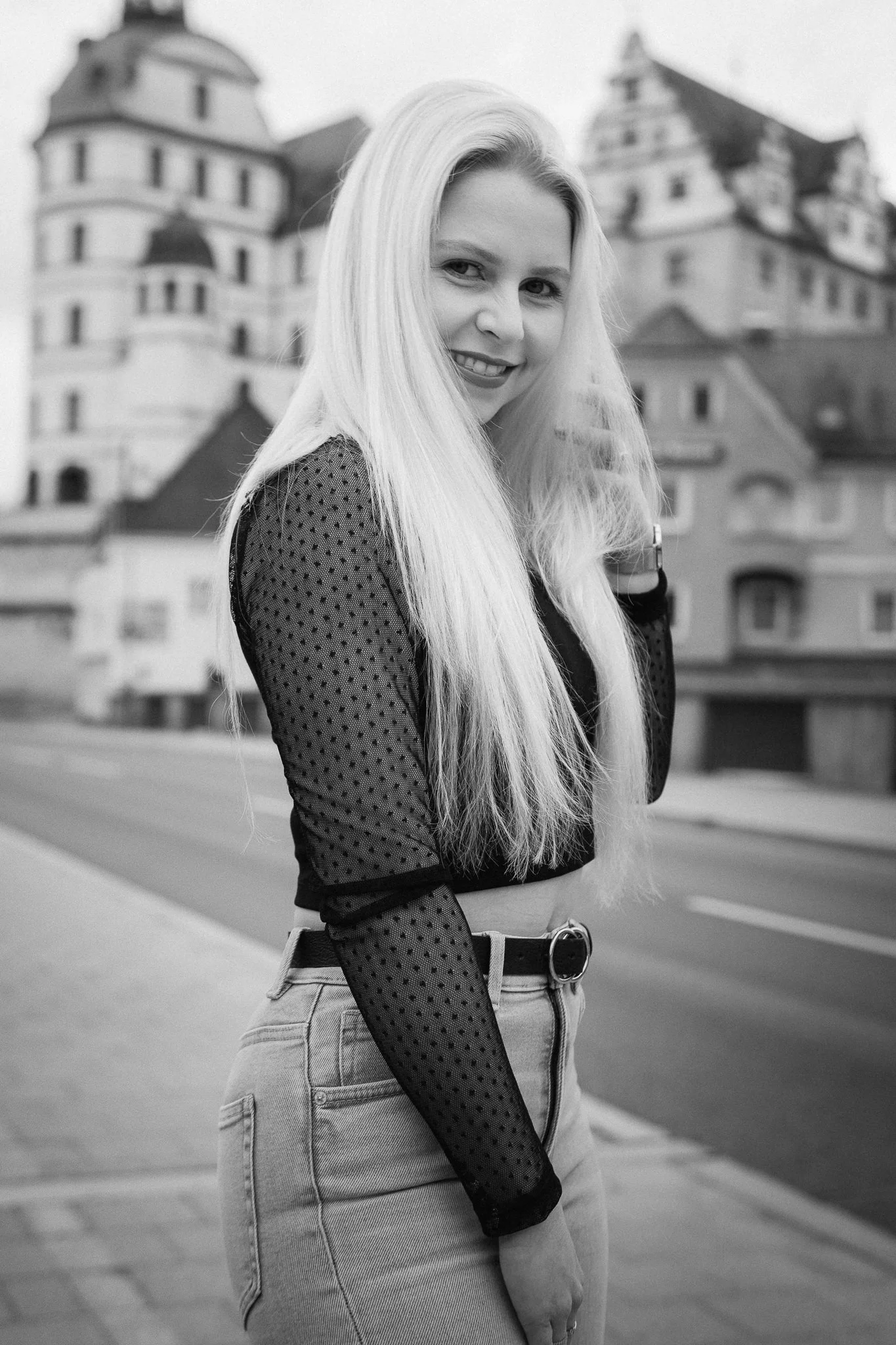 Junge Frau mit langen blondem Haar trägt schwarzen Hosenträger-Top, Jeans und Uhr, steht vor historischen Gebäuden in einer Stadt im Black-and-White-Foto.
