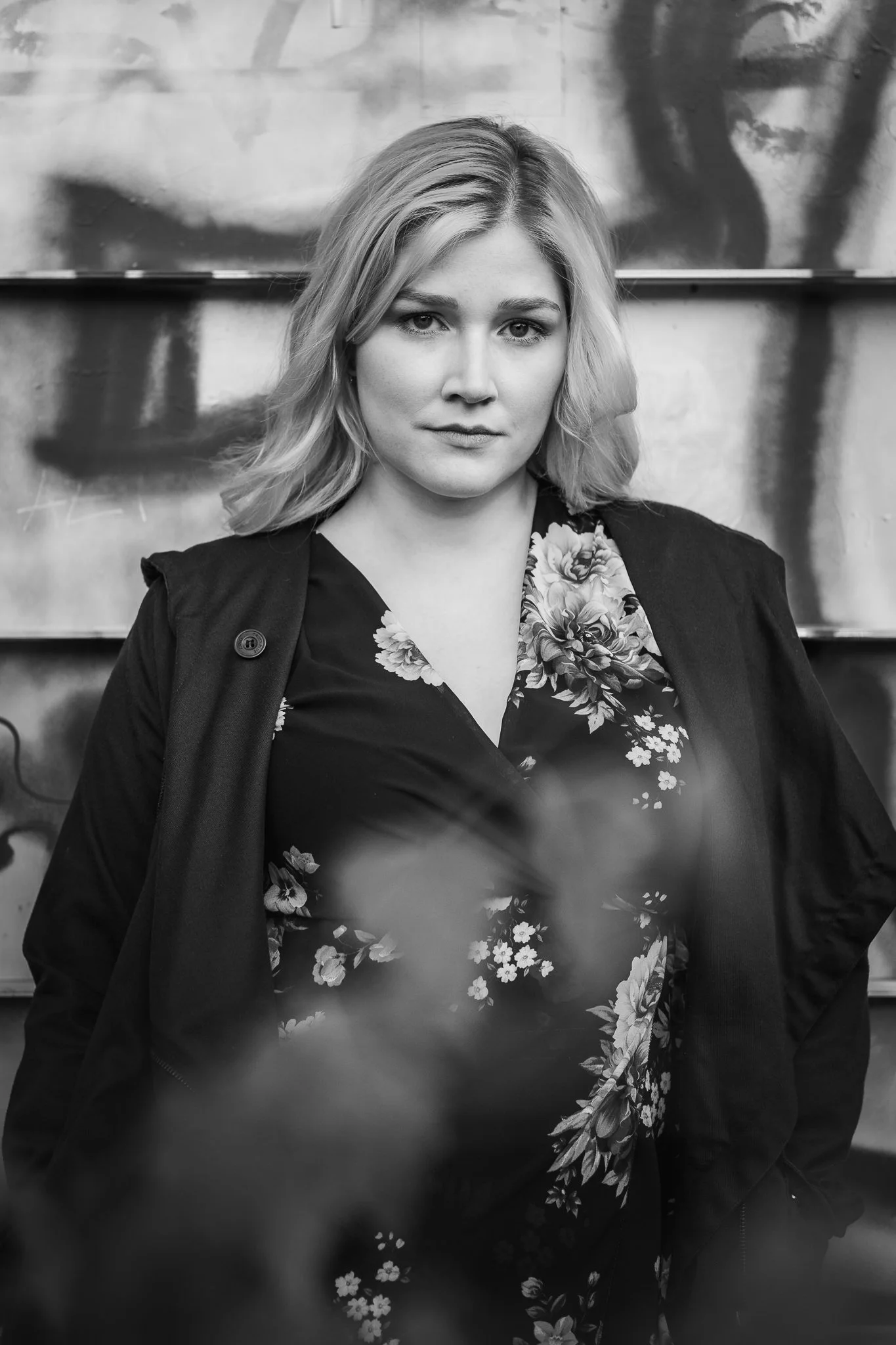 Schwarz-weiß-Fotografie einer jungen Frau mit Schulterlänge Haaren, die eine dunkle Bluse mit floralem Muster trägt und vor einer Wand mit Graffiti steht.