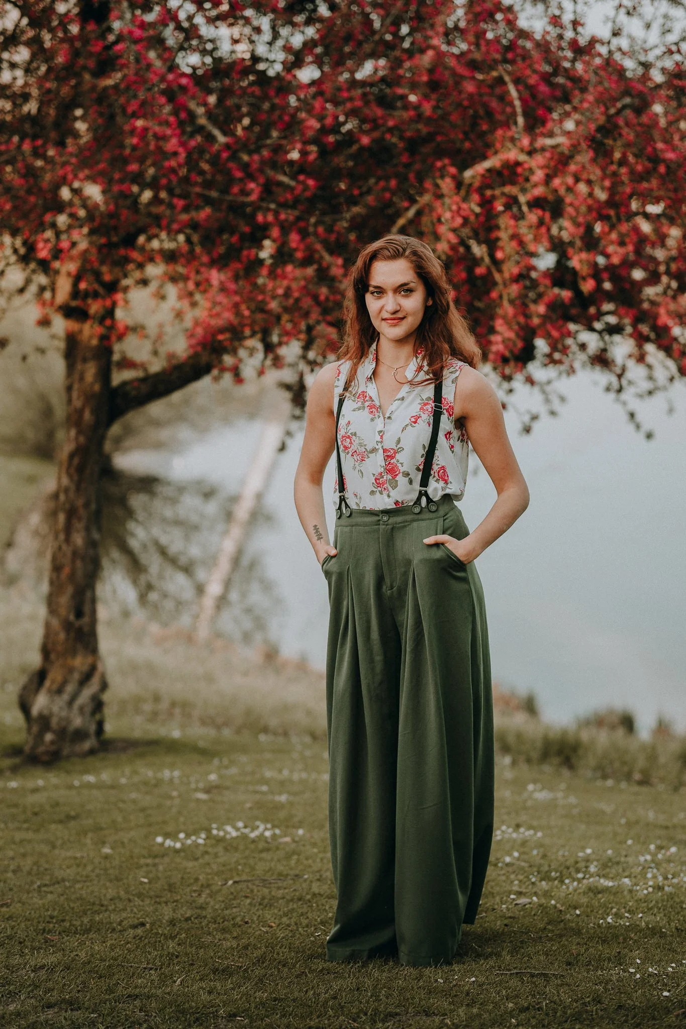 Junge Frau mit braunen, gewellten Haaren trägt ein ärmelloses, weißes Oberteil mit rosanen Blumenmuster, grüne weite Hosen und Suspenders, steht im Freien auf grünem Rasen vor einem Baum mit roten Blüten, im Hintergrund ein See.
