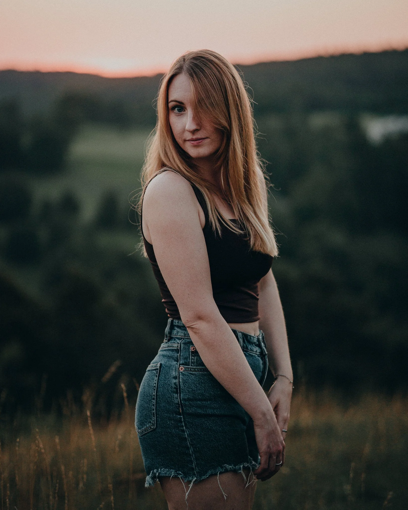 Eine junge Frau mit rotbraunen Haaren steht an einem Abend im Freien vor einer bewaldeten Hügelkette, trägt ein schwarzes Top und eine jeansshorts, blickt etwas zur Seite.
