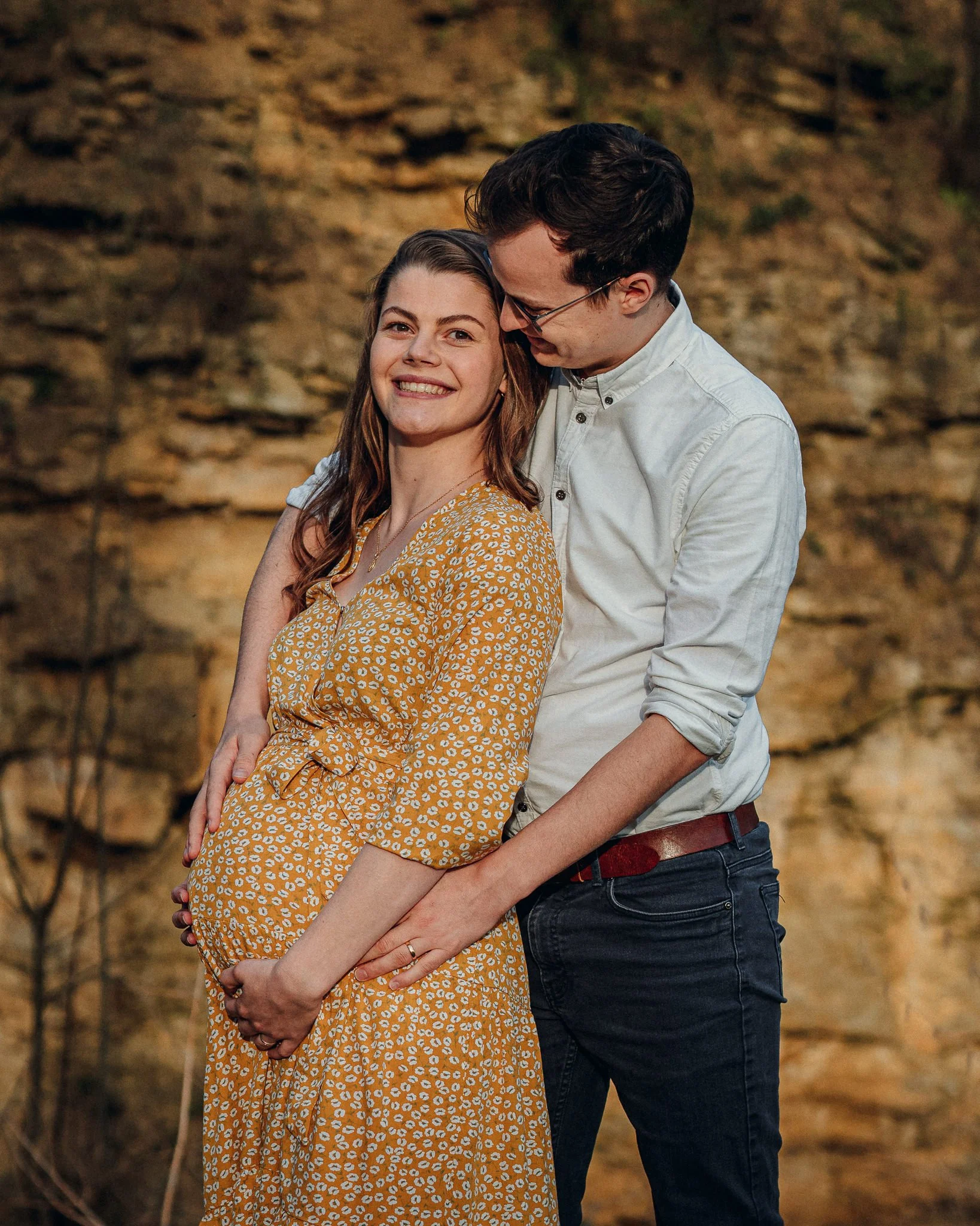 Ein schwangeres Paar steht vor einer Felswand, lächelt und schaut sich liebevoll an.