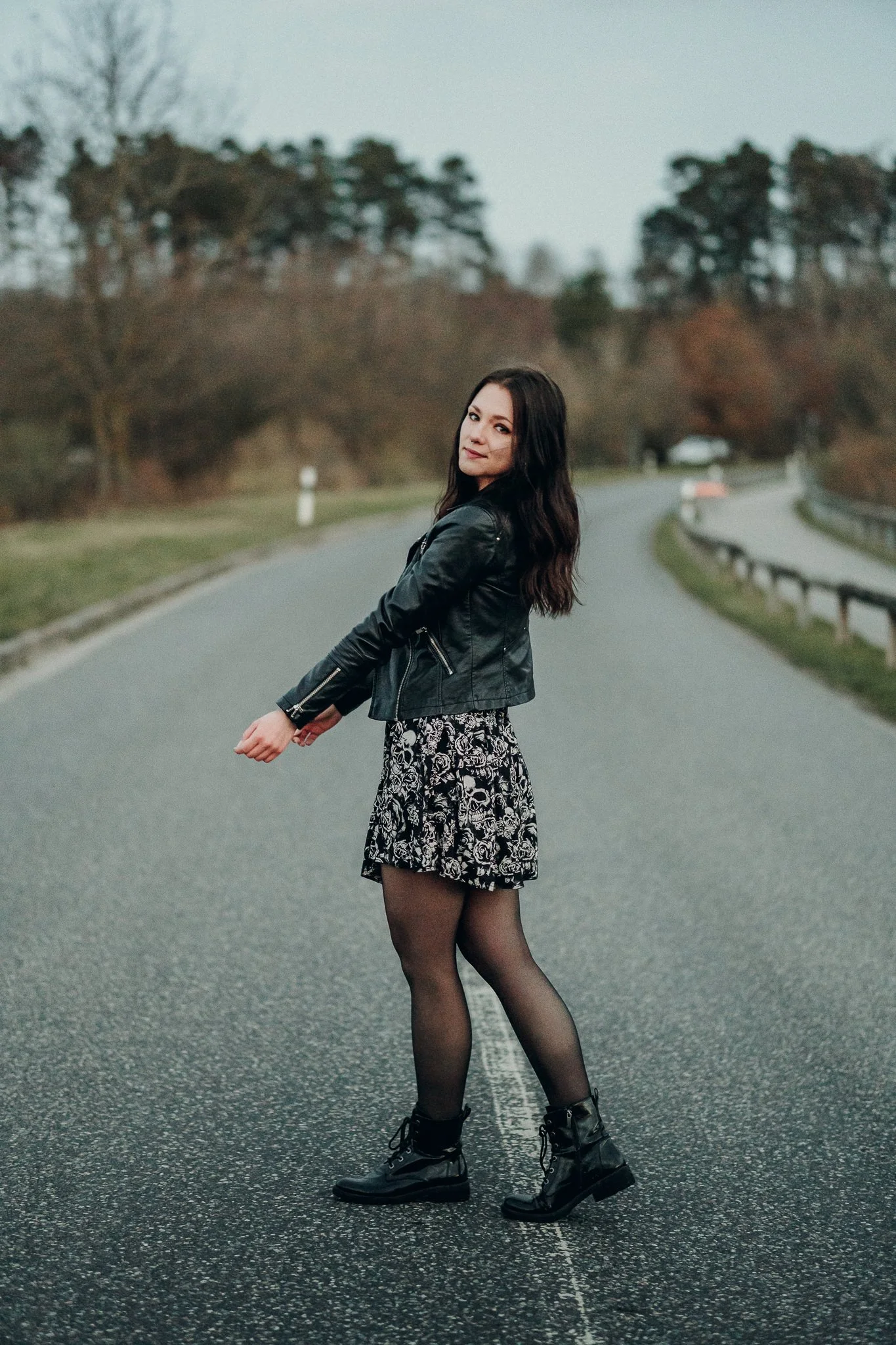 Junge Frau steht auf einer leeren Straße in einem Park, trägt schwarzen Lederjacke, floral gemusterten Rock, schwarze Strumpfhosen und schwarze Stiefel, mit dunklen, lockigen Haaren, im Hintergrund herbstliche Bäume.