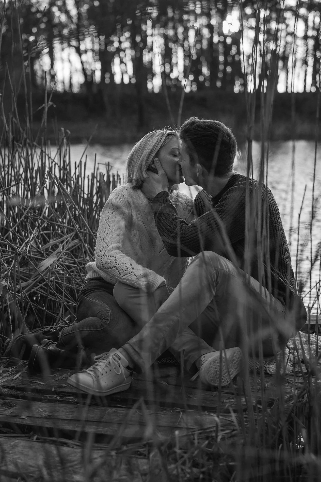 Ein Paar sitzt an einem See, küsst sich und umarmt sich, umgeben von Schilf und Bäumen im Hintergrund, in Schwarzweiß.