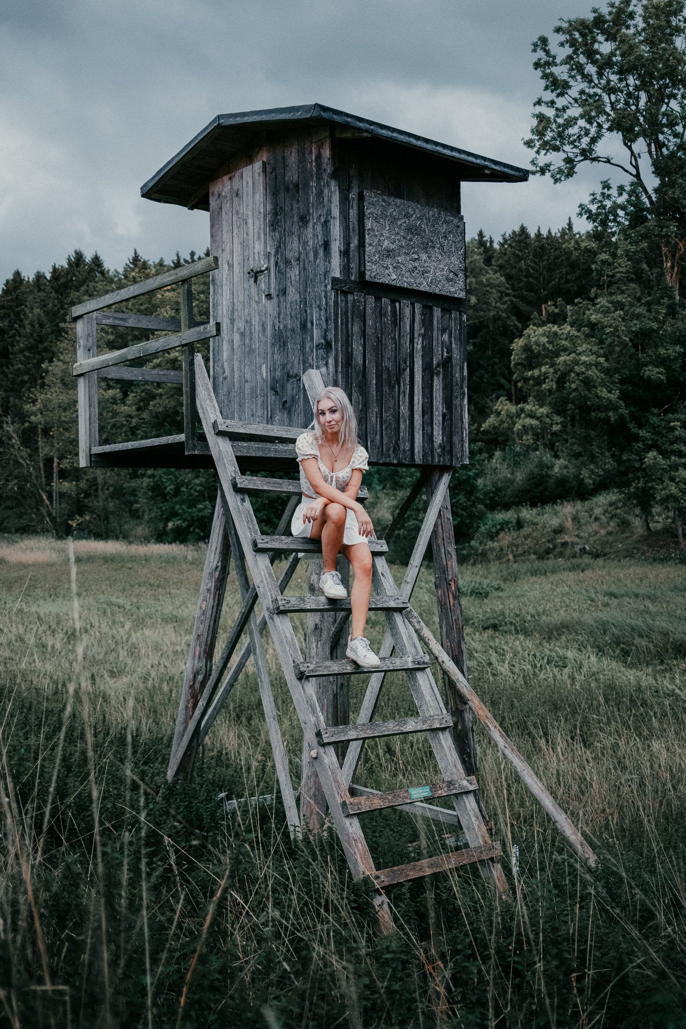 Junge Frau sitzt auf einer hölzernen Leiter vor einem Baumhaus in einem grasbewachsenen Feld, umgeben von Bäumen, bei wolkigem Himmel.