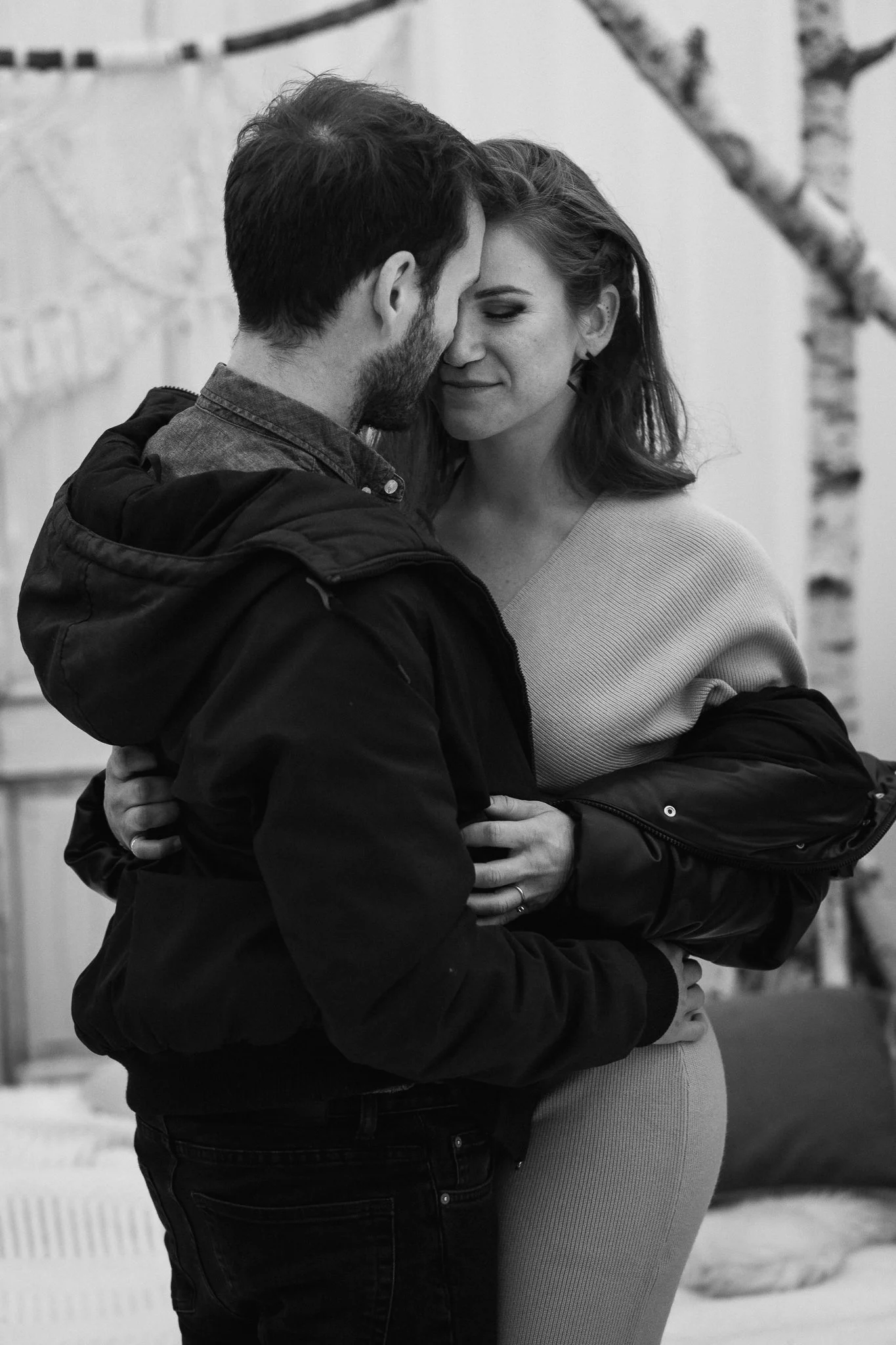 Ein Mann und eine Frau stehen eng umarmt, die Stirn aneinander, in einem Raum mit einem Baum im Hintergrund, in einem schwarzen und weißen Foto.