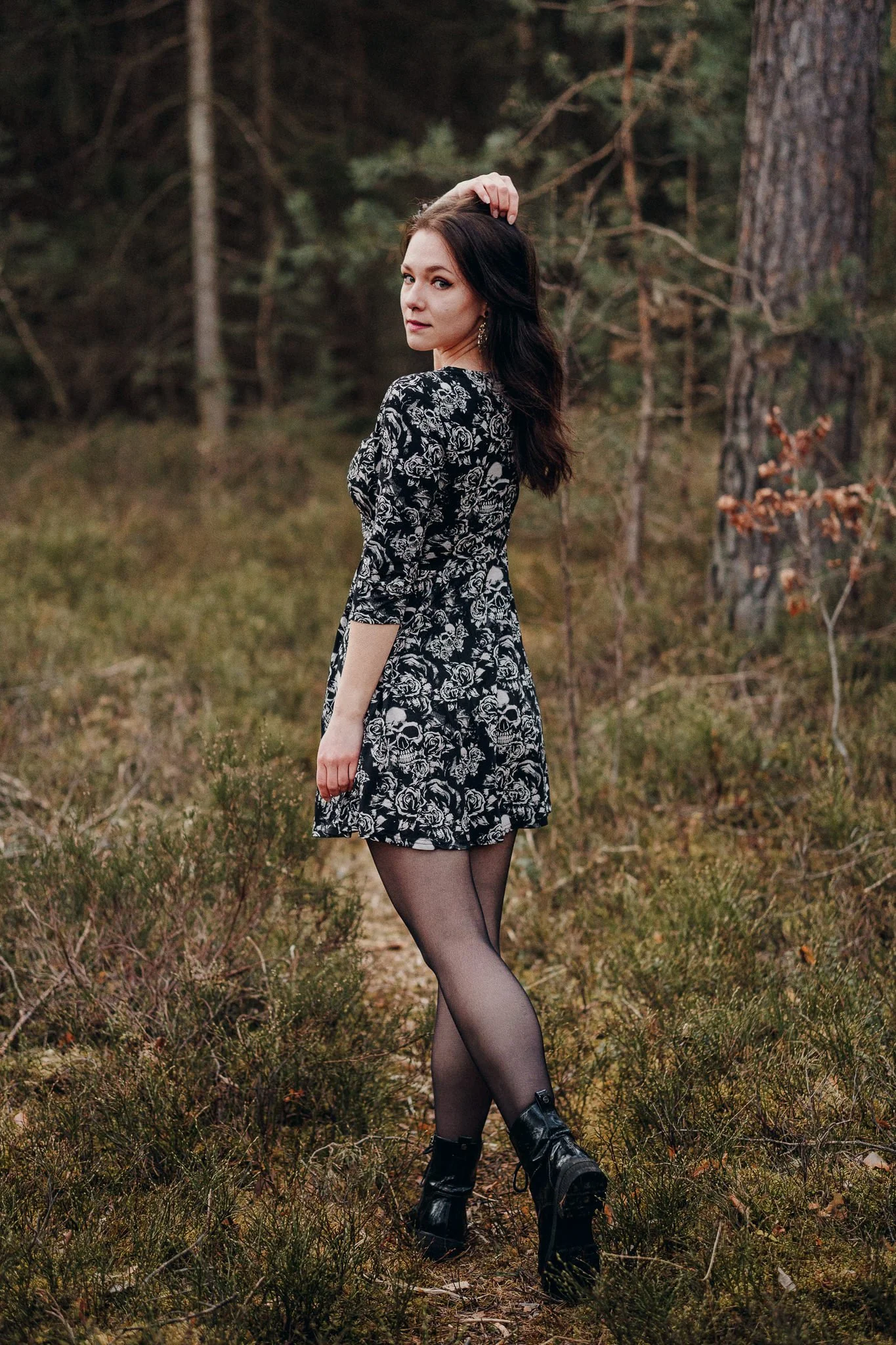 Eine junge Frau in einem schwarzen Kleid mit weißen Rosendetails und Totenkopfmustern steht im Wald mit Büschen und Bäumen im Hintergrund.
