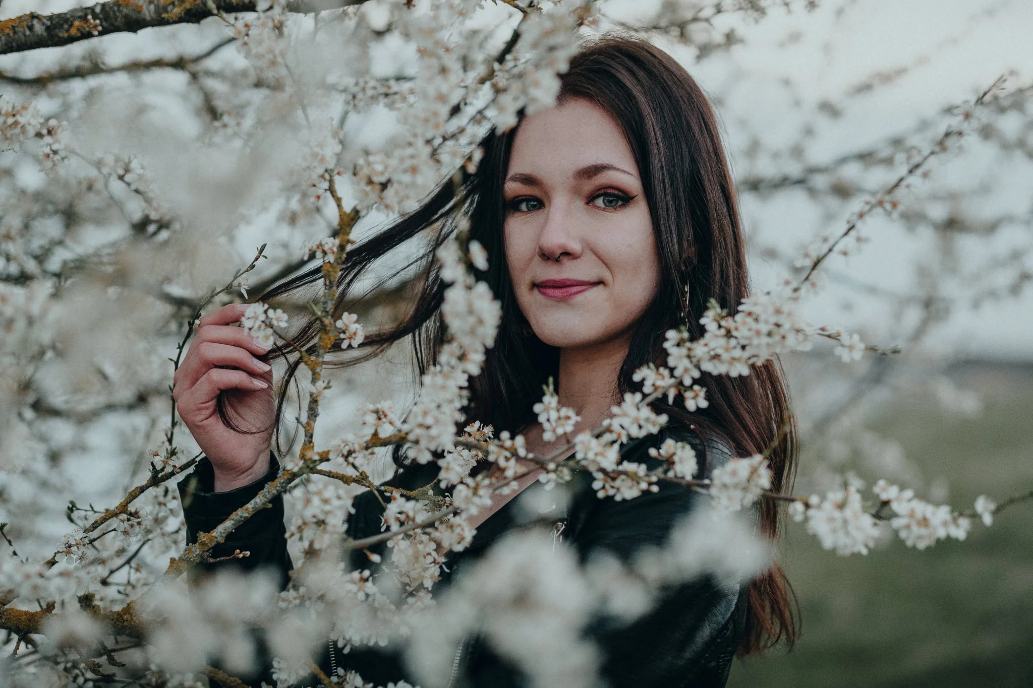 Eine junge Frau mit dunklem Haar und Make-up steht zwischen blühenden weißen Kirschblüten und lächelt leicht.