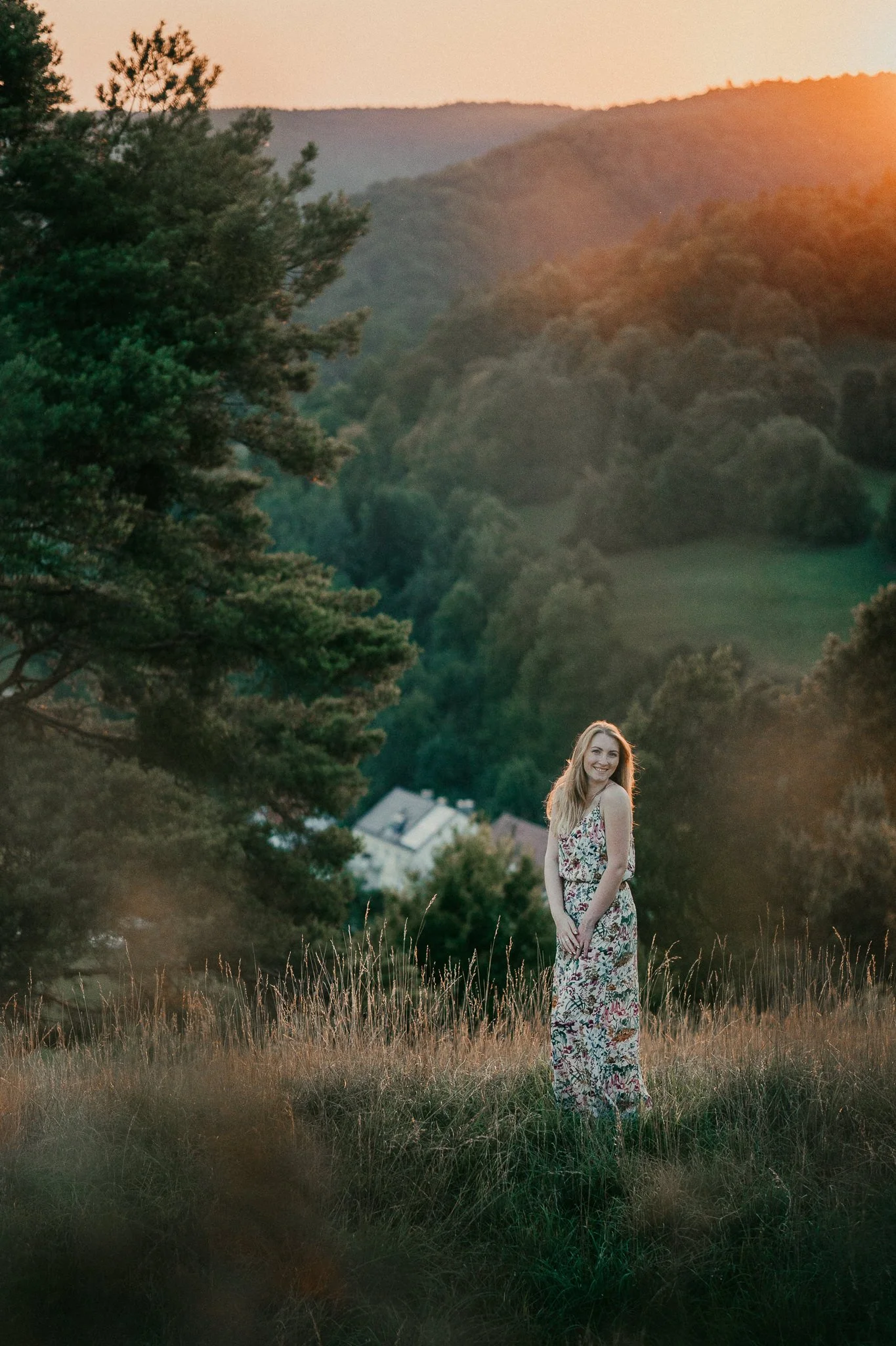Eine lachende junge Frau in einem langärmligen, bunten Kleid steht in einer Wiesenlandschaft bei Sonnenuntergang, mit Bäumen und Hügel im Hintergrund.