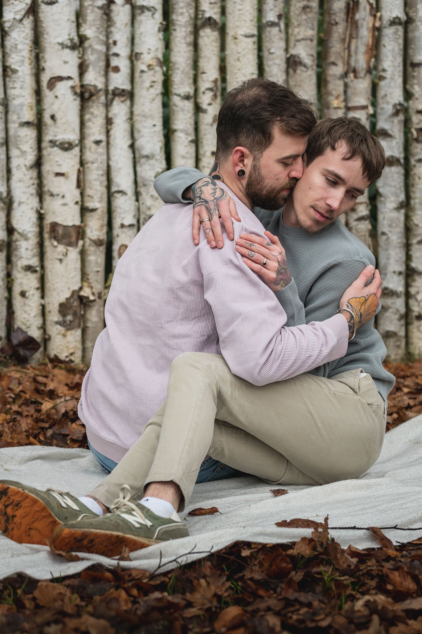 Zwei Männer umarmen sich auf einer Decke im Wald, vor einer Birkenholz-Wand, umgeben von Herbstlaub.