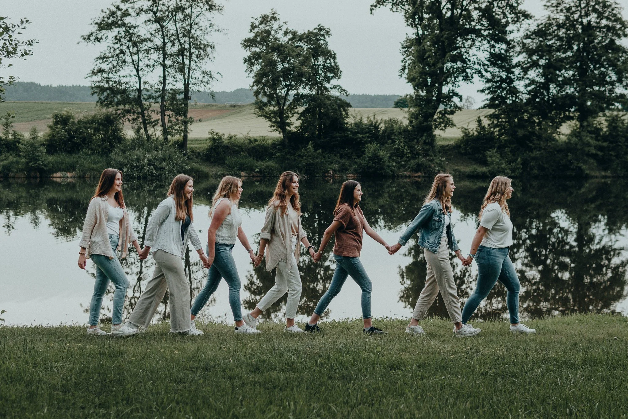 Sieben junge Frauen halten sich an den Händen und gehen an einem Teich in einer grünen, ländlichen Umgebung.