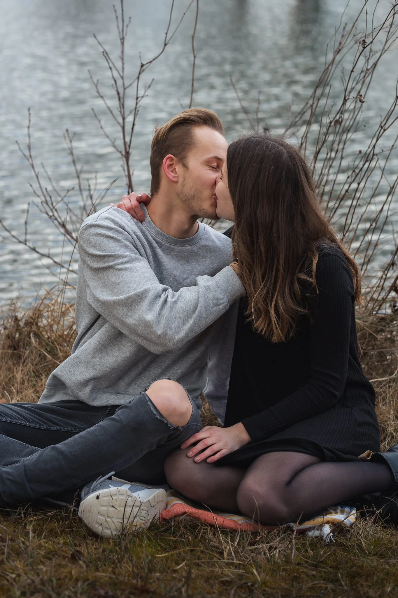 Ein junges Paar sitzt in der Natur am Wasser und küsst sich.