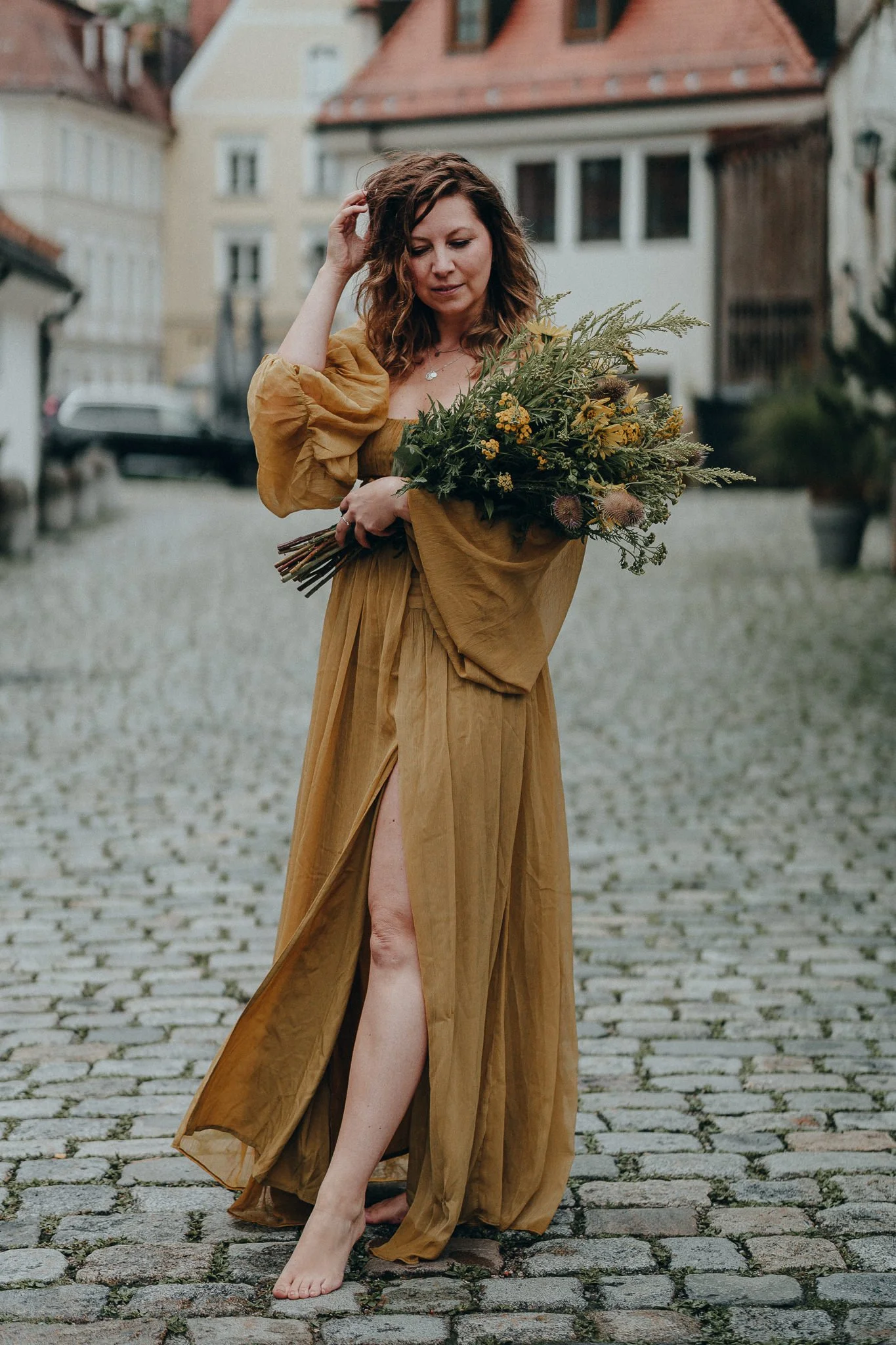 Frau in einem goldfarbenen Kleid auf einer Kopfsteinpflasterstraße, die einen Blumenstrauß hält, mit mittelalterlichen Gebäuden im Hintergrund.