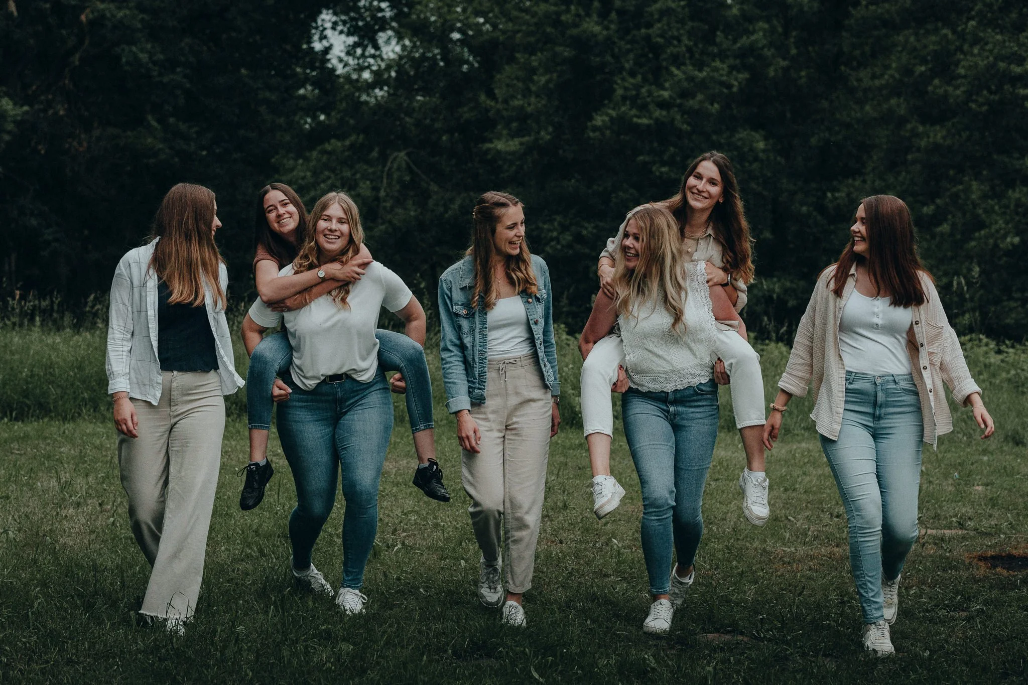 Sieben junge Frauen im Park, zwei tragen andere auf den Rücken, beim Spaß und Lachen.