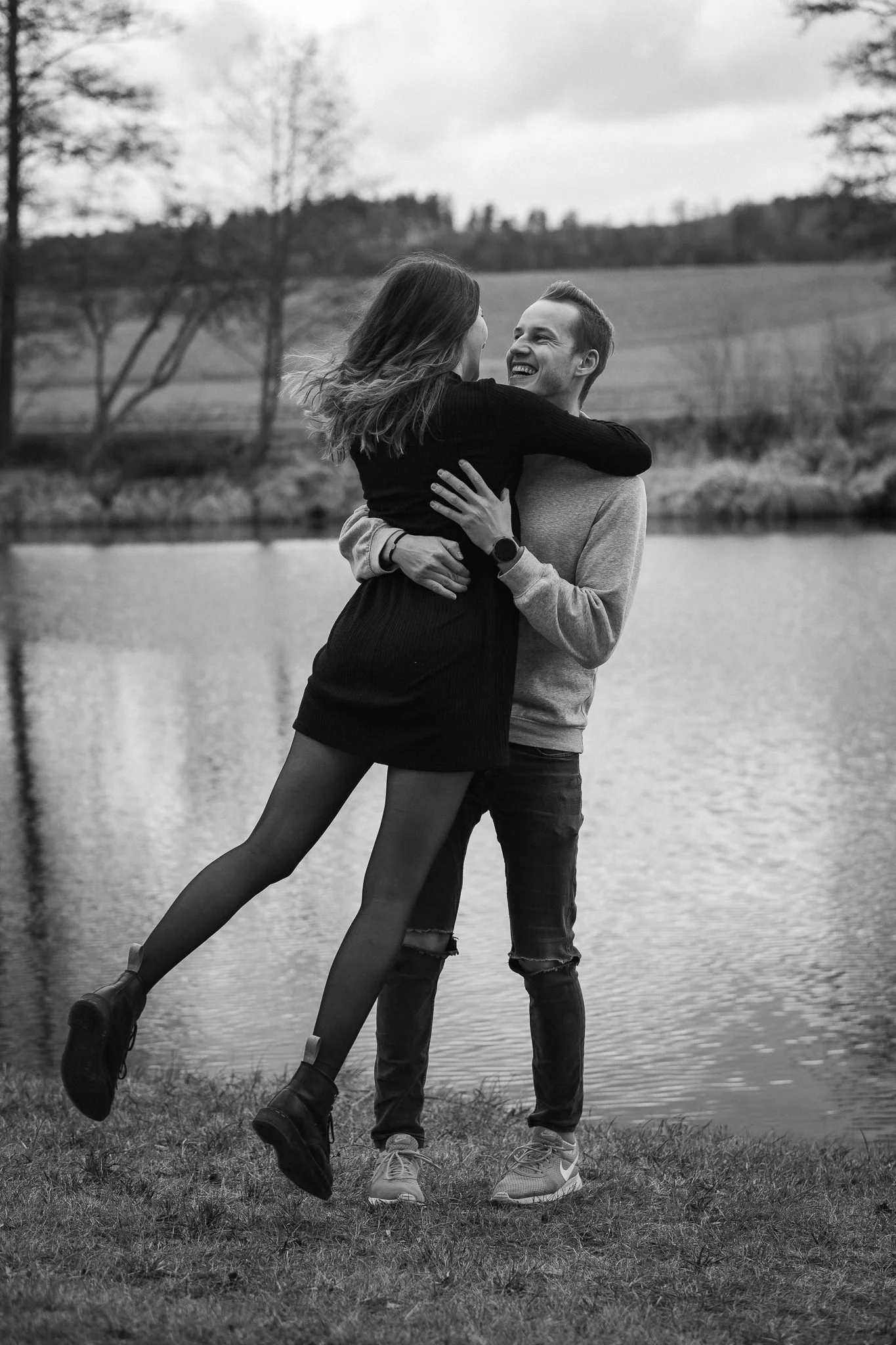 Ein lachendes Paar umarmt sich am Ufer eines Sees im Freien, umgeben von Bäumen und Wiesen, im schwarzen und weißen Foto.