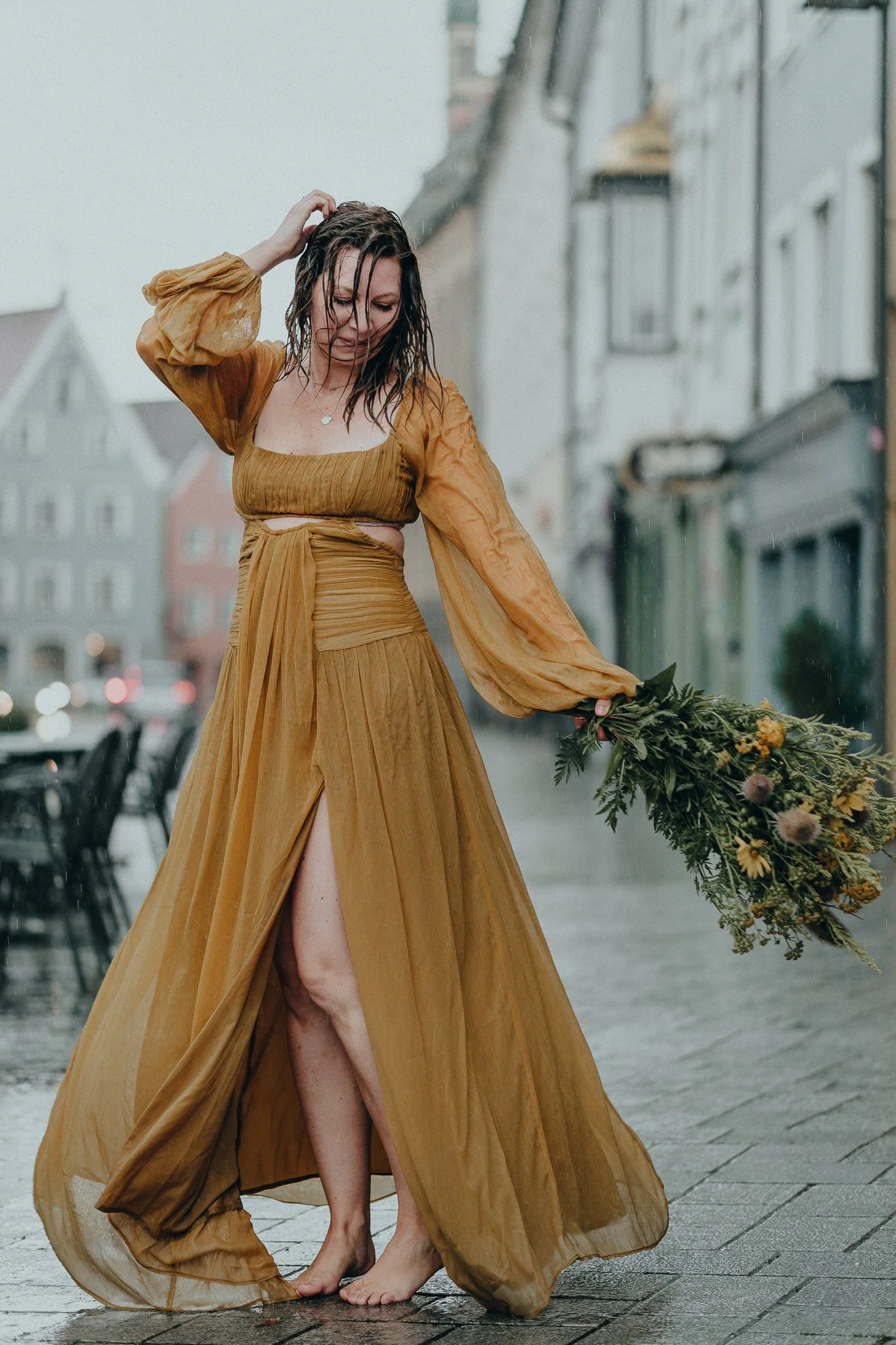 Eine Frau in einem goldbraunen, bodenlangen Kleid steht barfuß im Regen auf einer Straße, hält einen Blumenstrauß und scheint den Regen zu genießen.