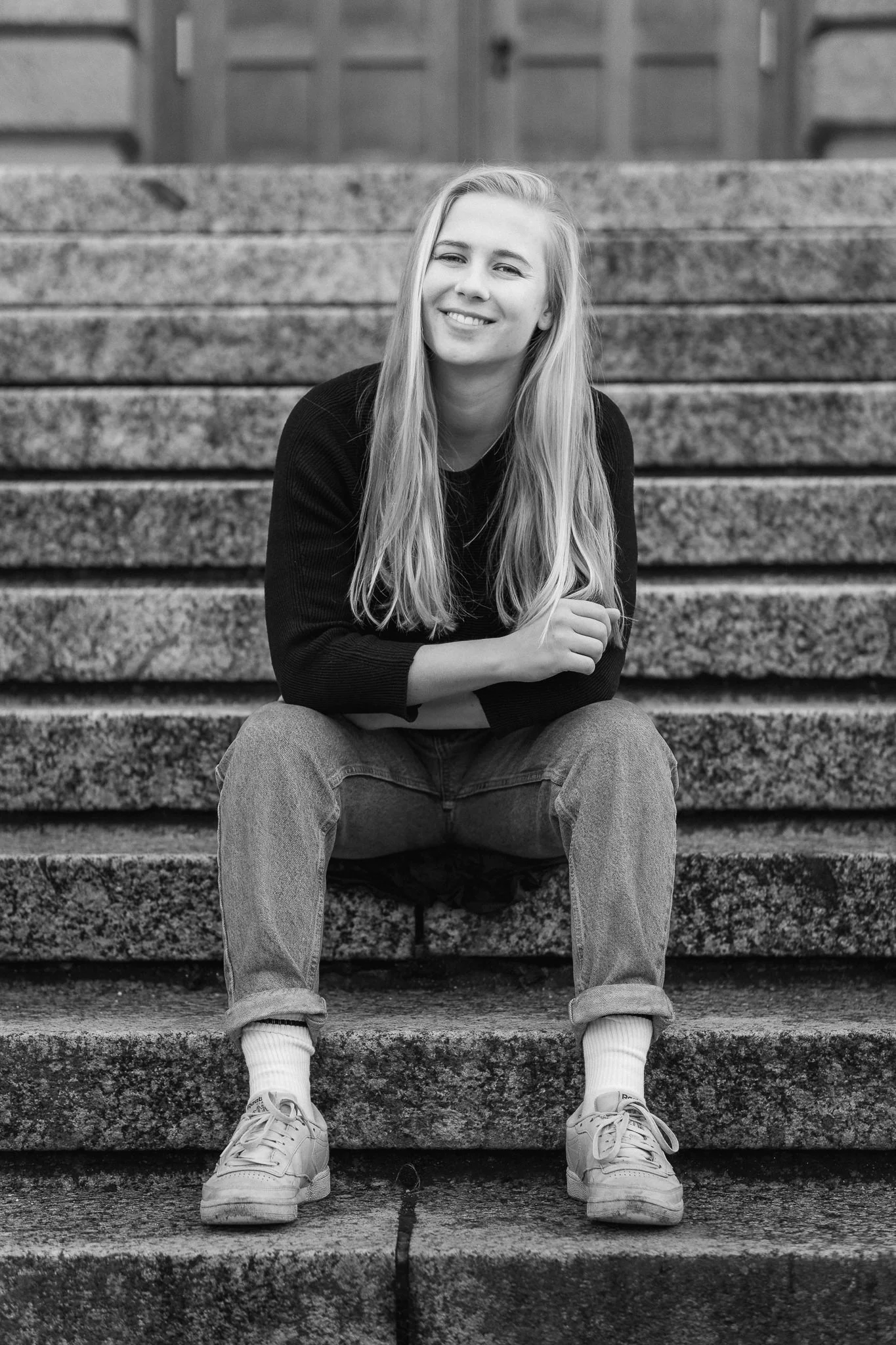 Schwarz-Weiß-Foto einer lächelnden jungen Frau mit langen Haaren, sitzend auf Steintreppen vor einem Holztor, gekleidet in eine schwarze Jacke, Jeans, weiße Socken und Turnschuhe.