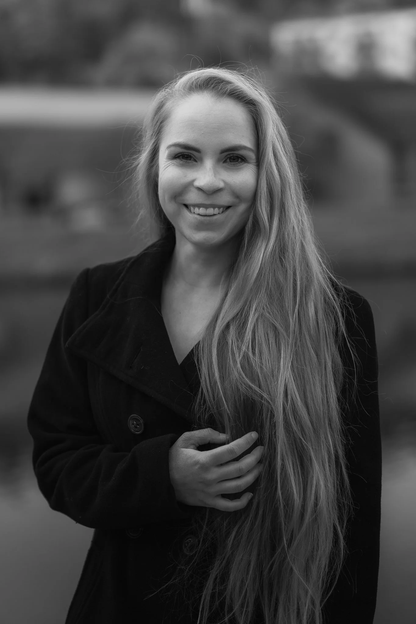 Lächelnde Frau mit langen Haaren vor unscharpem ländlichem Hintergrund, schwarz-weiß aufgenommen.