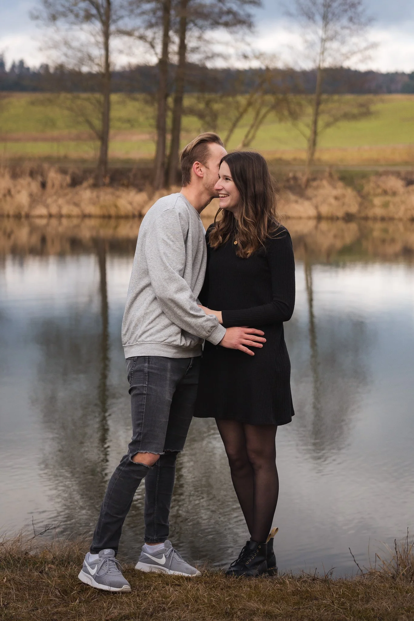 Ein verliebtes Paar steht an einem See, umgeben von Bäumen, bei Sonnenuntergang, lächelnd und sich berührend.