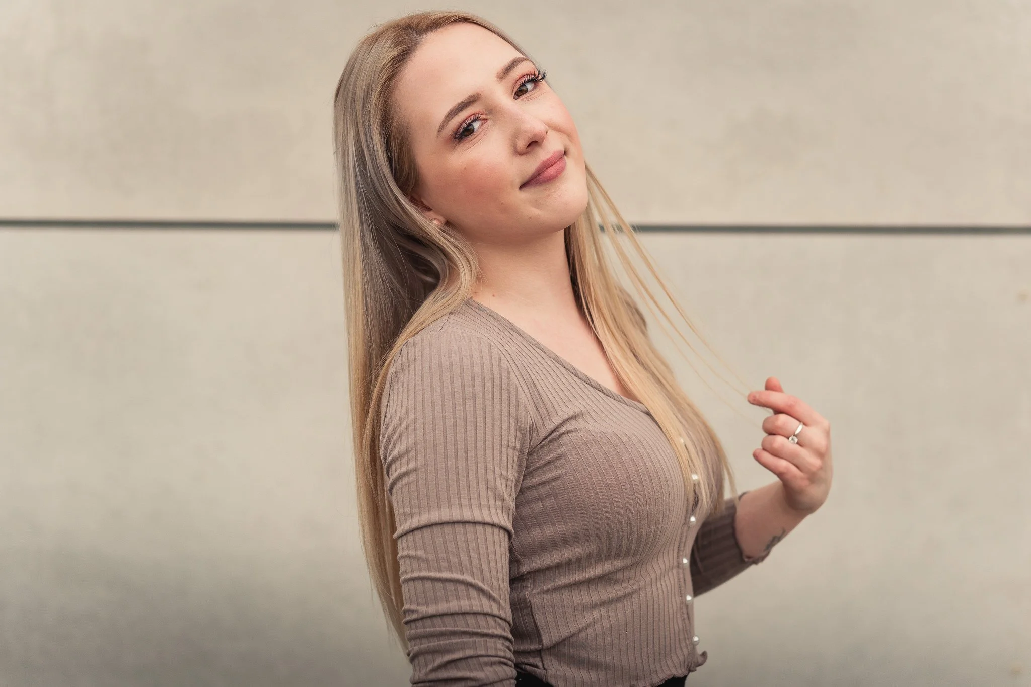 Junge Frau mit langem blondem Haar, trägt einen braunen Pullover, steht vor einer hellen Wand, lächelt leicht und streicht sich eine Strähne.