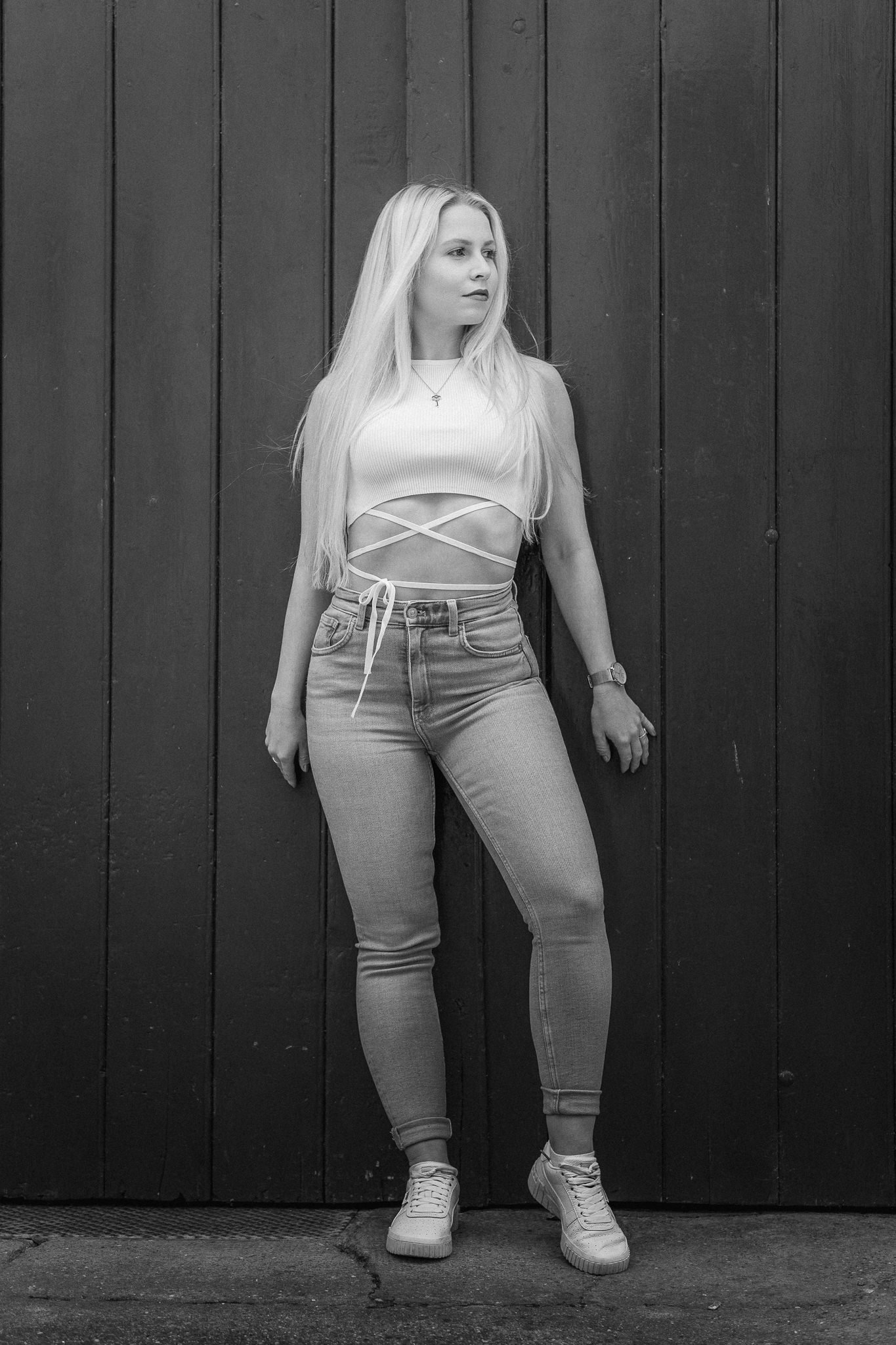 Junge Frau mit langem blondem Haar steht vor einer dunklen Holztür, trägt einen Crop-Top und Jeans, in Schwarz-Weiß-Fotografie.