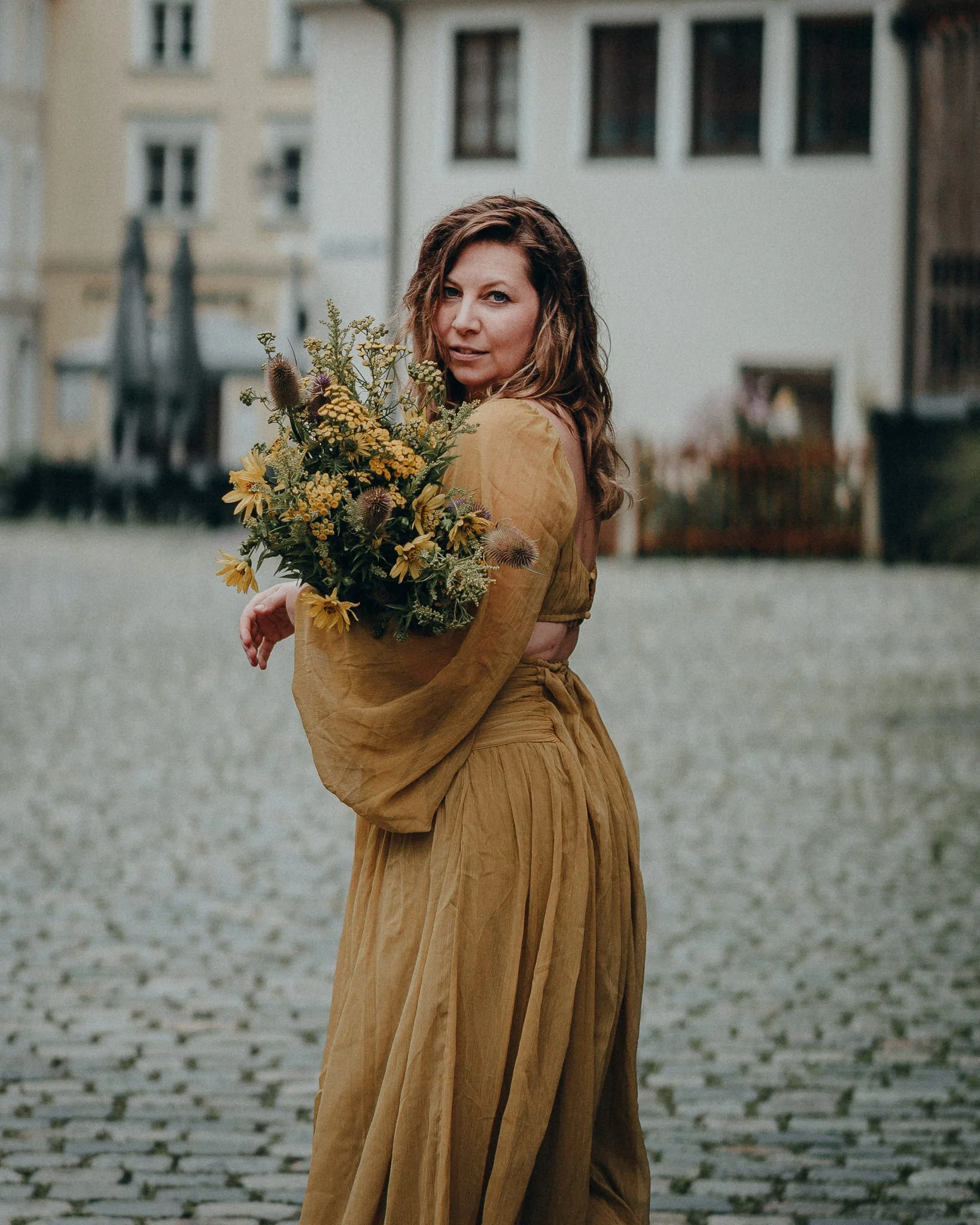 Eine Frau in braunen Kleidern hält einen Blumenstrauß auf einer Kopfsteinpflasterstraße, im Hintergrund Gebäude. Sie blickt zur Kamera.