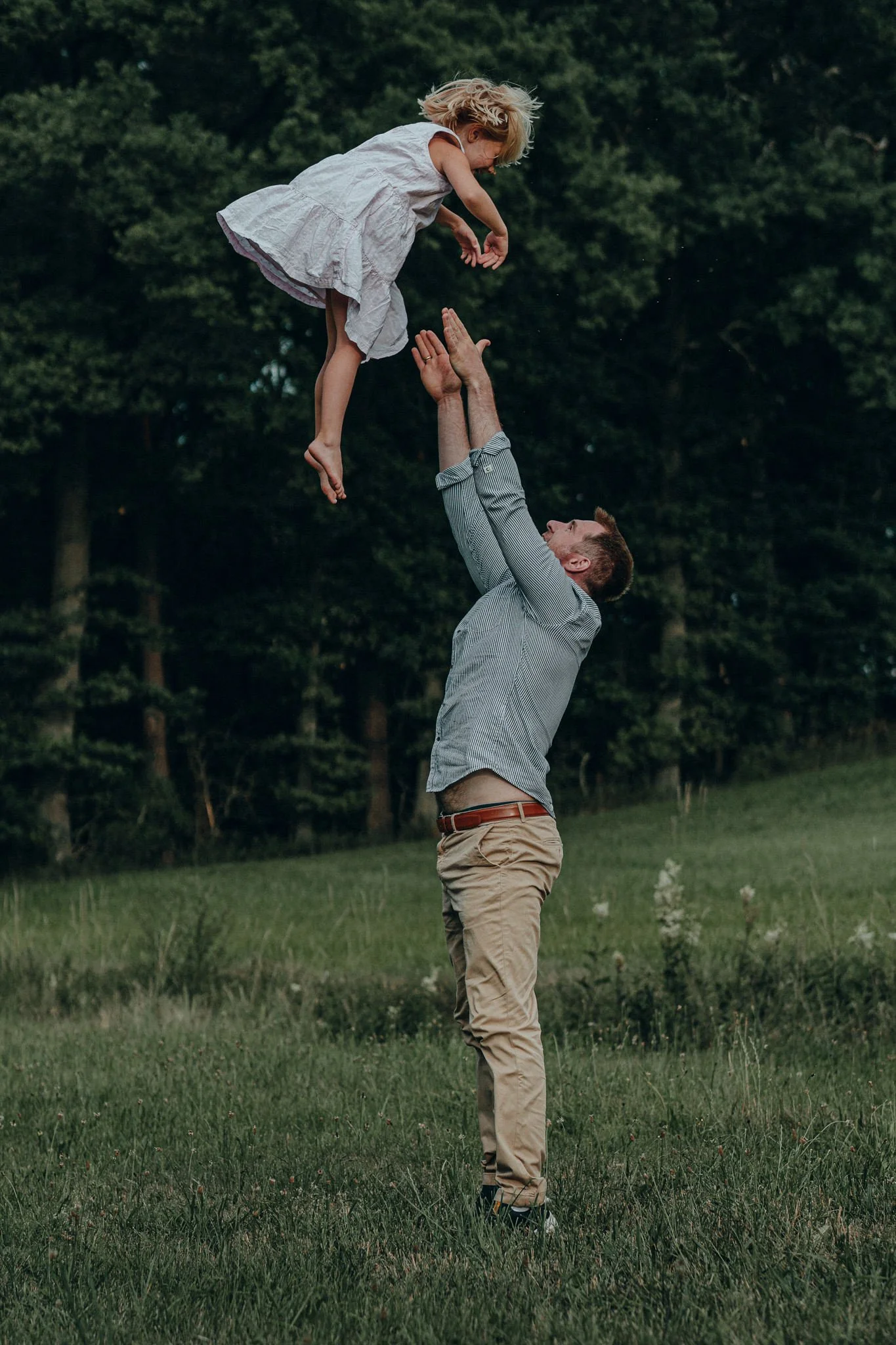 Vater wirft Tochter in der Natur in die Luft, beide lachen, im Hintergrund Bäume und Wiese.