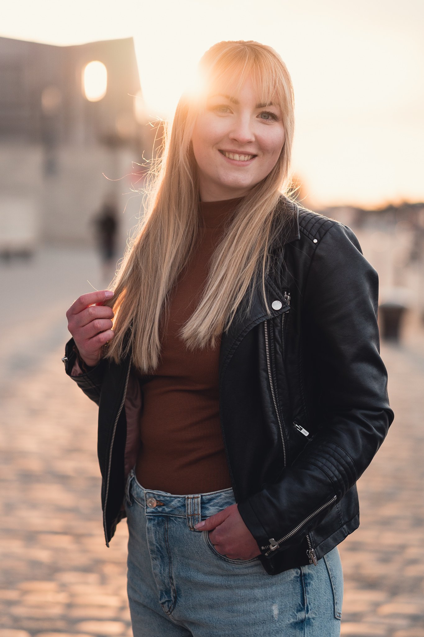 Junge Frau mit langblondem Haar, schwarzer Lederjacke, braunem Oberteil und blauen Jeans, lächelnd bei Sonnenuntergang am Wasser.