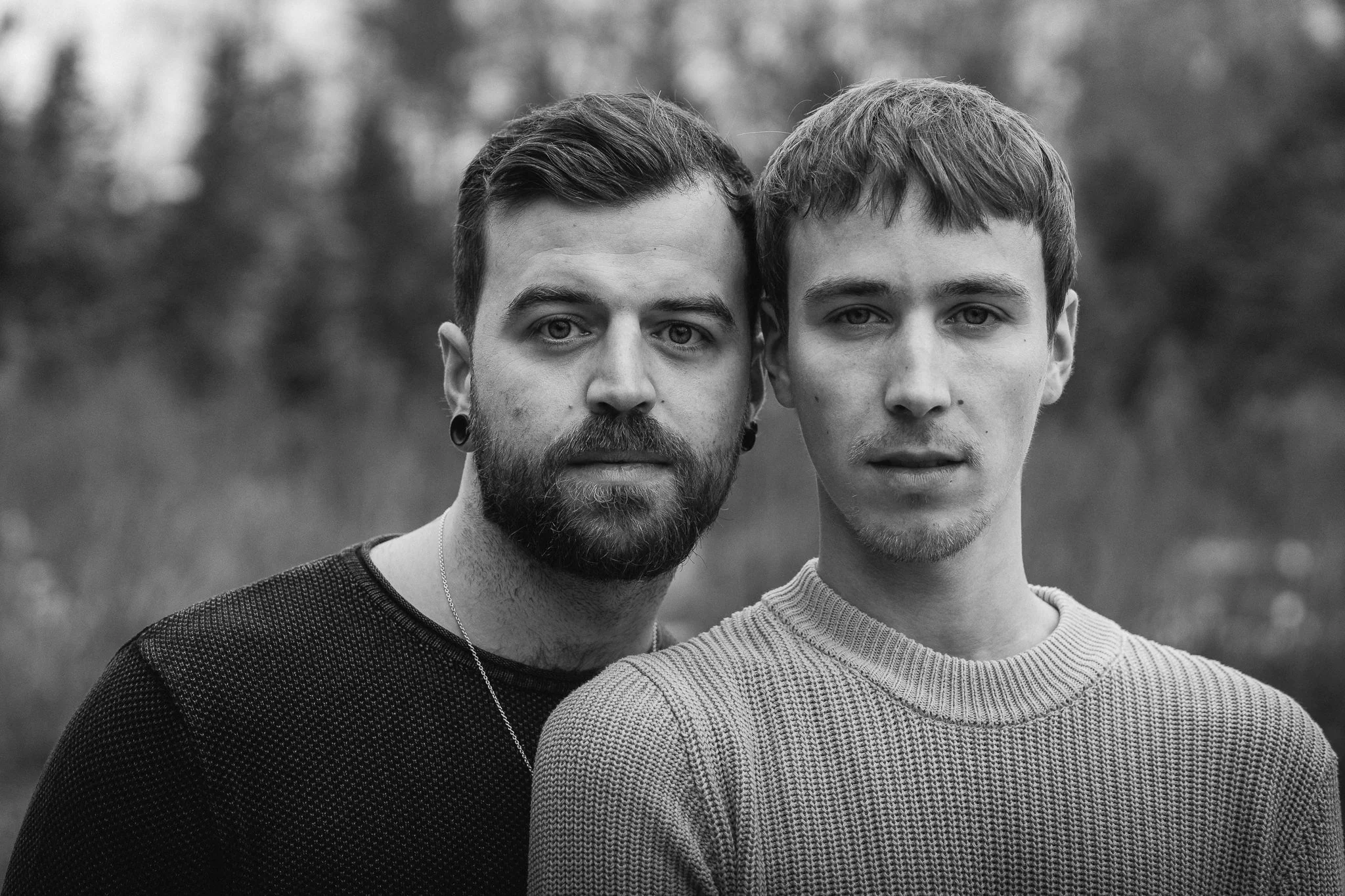 Zwei junge Männer stehen nahe beieinander im Freien, beide schauen direkt in die Kamera, im Hintergrund unscharfe Natur, Schwarz-Weiß-Foto.
