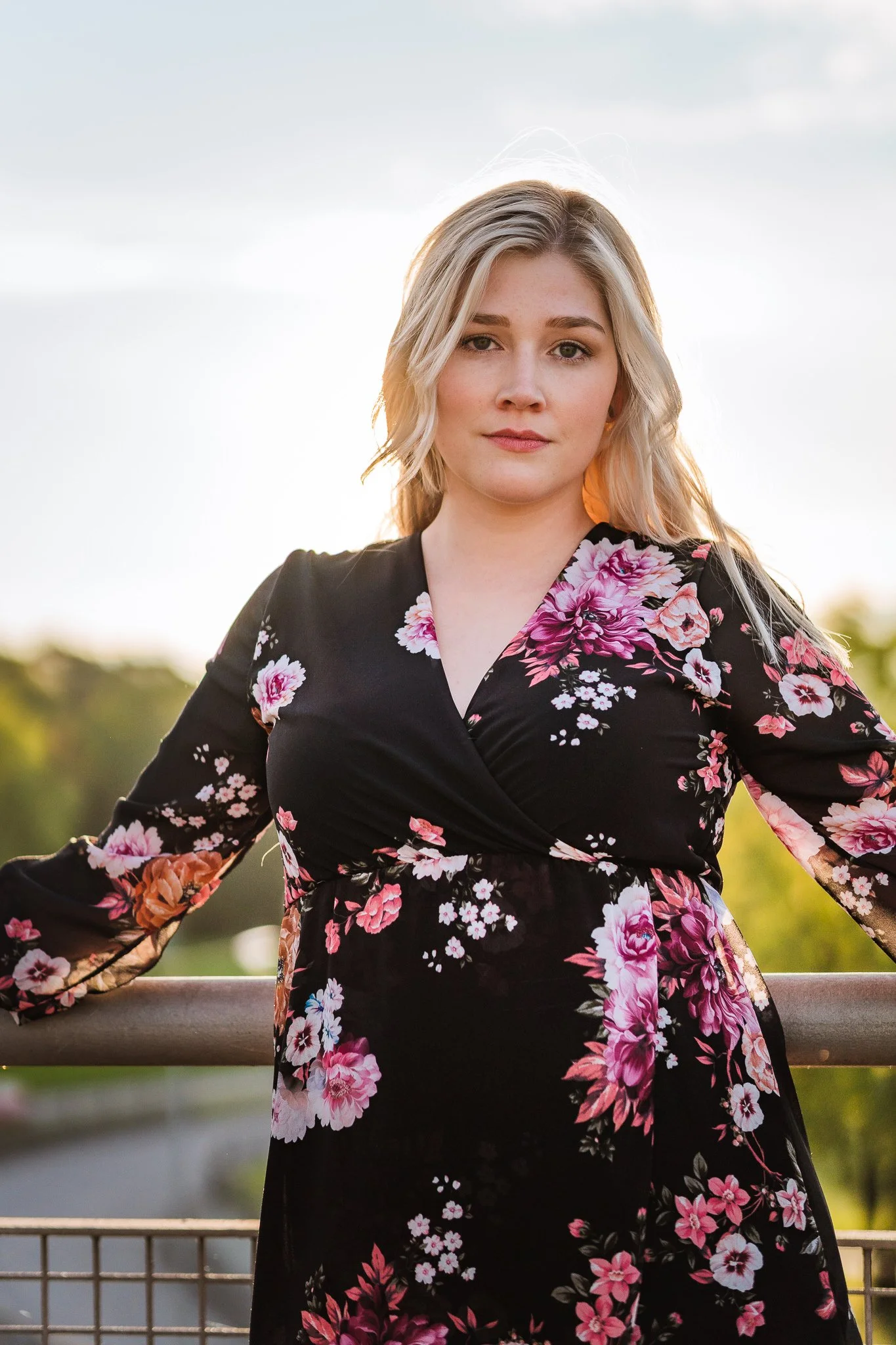 Eine junge Frau mit langen blonden Haaren trägt ein schwarzes Kleid mit pink-weißen Blumenmuster und steht vor einer natürlichen Hintergrundkulisse im Freien.