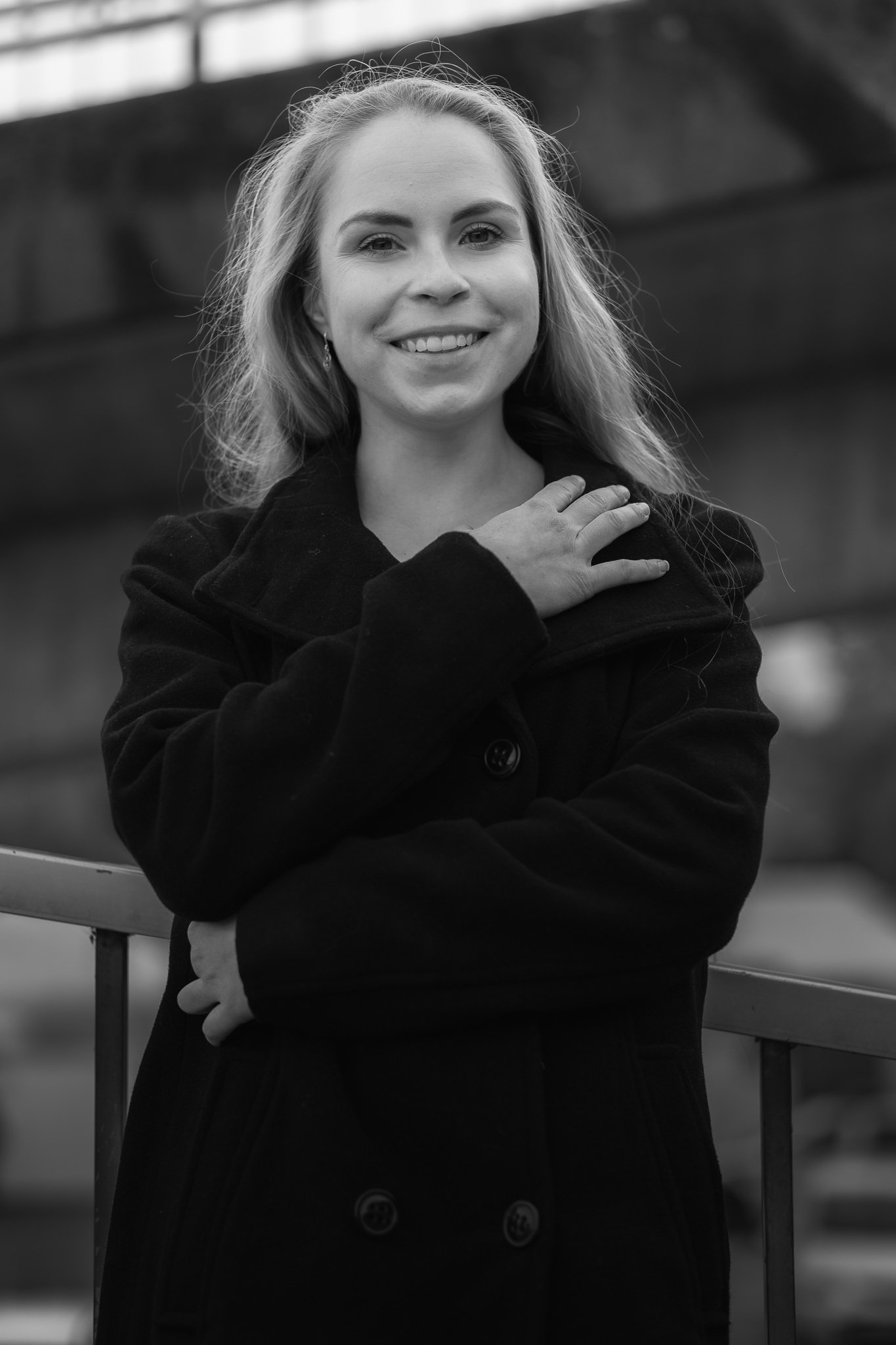 Schwarz-weiß-Fotografie einer lachenden Frau mit blondem Haar, die eine schwarze Jacke trägt, die an einem Geländer lehnt.