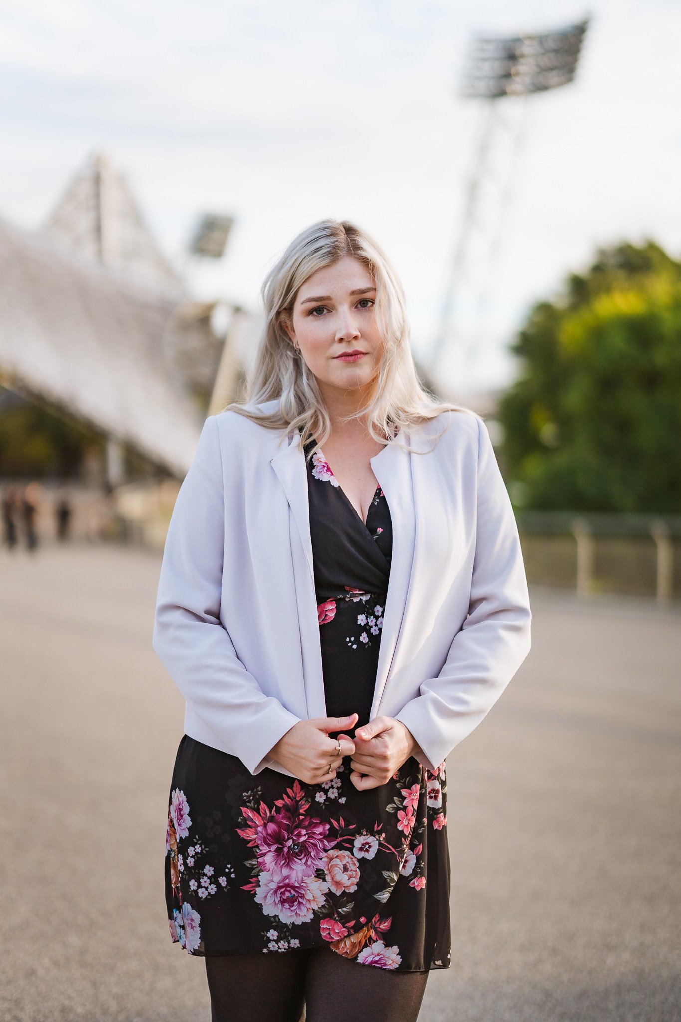 Junge blonde Frau steht draußen vor einem Sportstadion mit Flutlichtmasten, trägt schwarze Kleidung mit Blumenmuster, weiße Jacke, zeigt nach vorne.