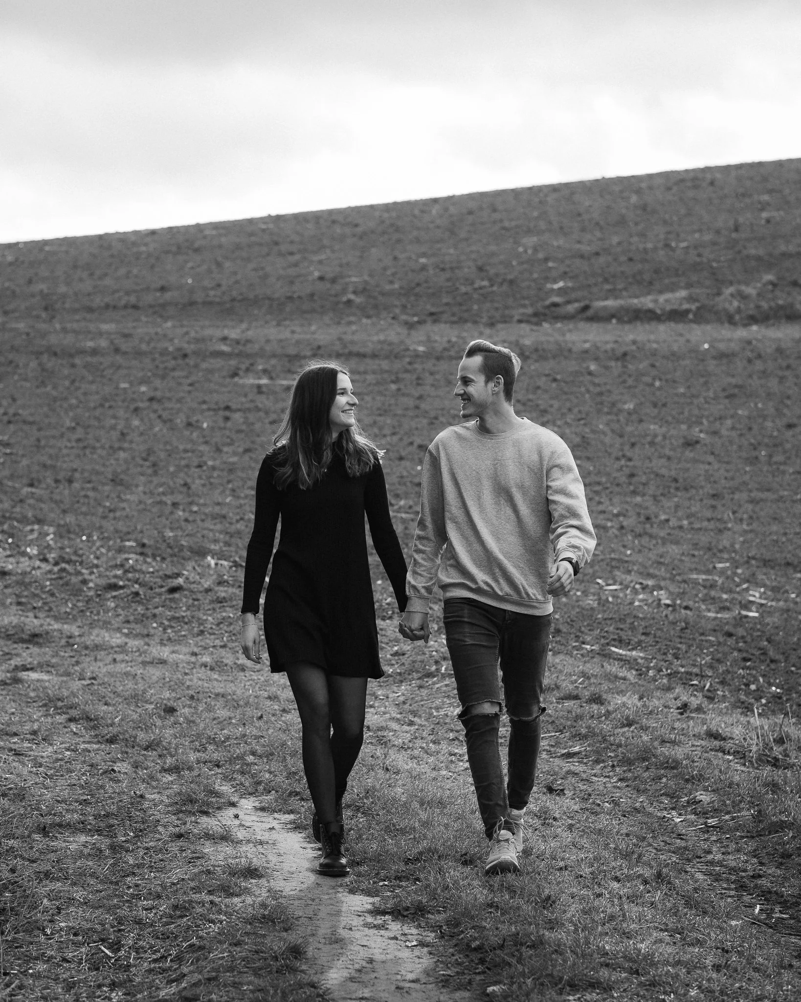 Ein junges Paar läuft Hand in Hand auf einem Feld, sie lächeln sich an, im Hintergrund ein Hügel, Schwarz-Weiß-Foto.