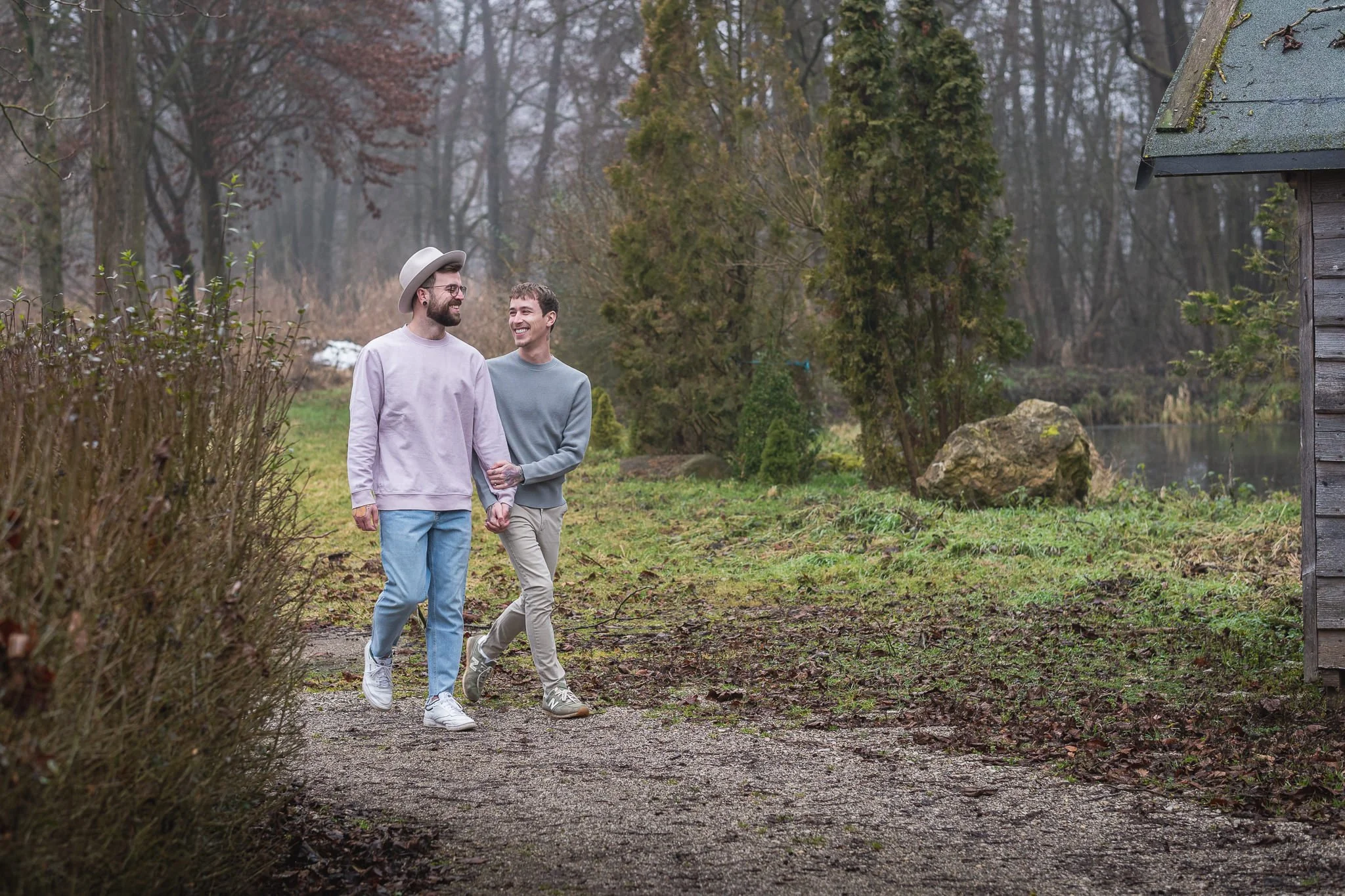 Zwei Männer laufen Hand in Hand auf einem Waldweg im Freien, umgeben von Büschen und Bäumen, bei bewölktem Himmel.