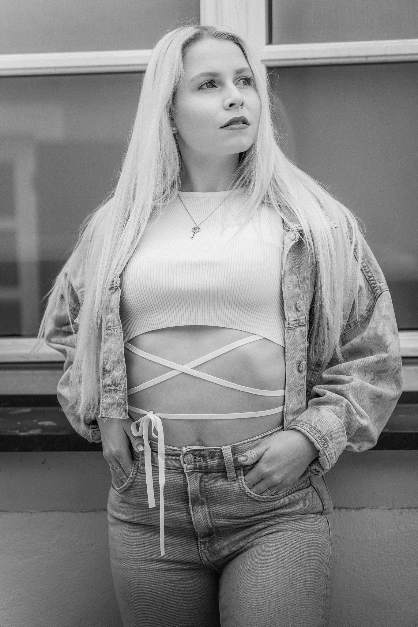 Schwarz-weiß-Foto einer jungen Frau mit langen, blonden Haaren, die in einem urbanen Umfeld steht. Sie trägt ein Crop-Top mit langen Bändern, die ihren Bauch umkreisen, eine Jeansjacke und Jeanshose, und schaut nach links.