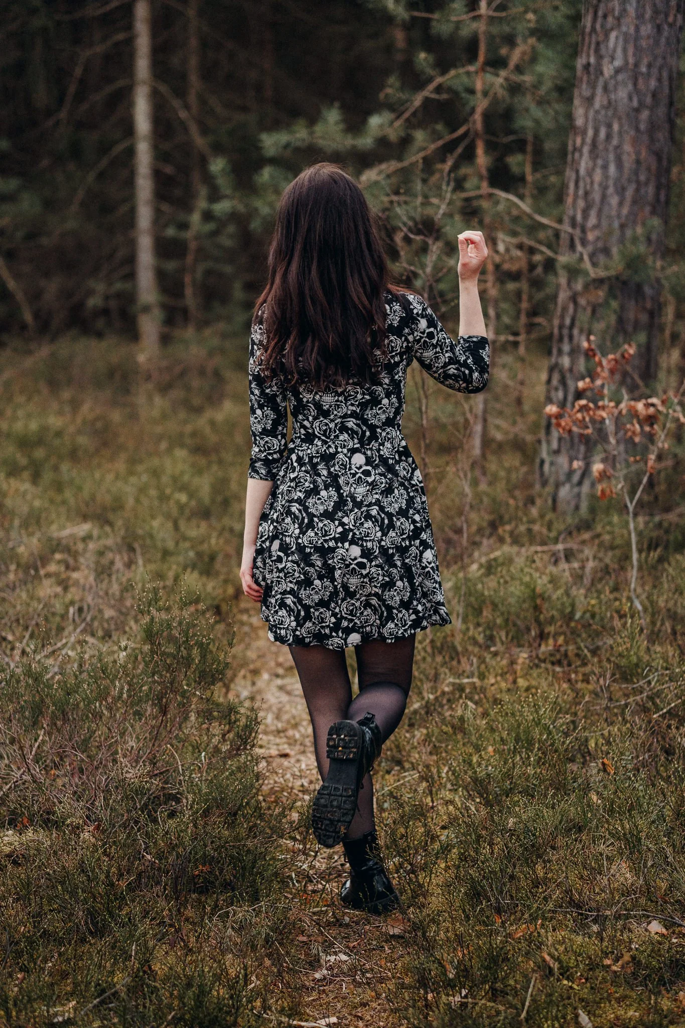 Frau mit dunklen Haaren läuft auf einem Waldweg, trägt ein schwarzes Kleid mit weißen Mustern, schwarze Strumpfhosen und Stiefel, Blick nach vorne gerichtet, in einer Naturumgebung mit Bäumen und Gras.