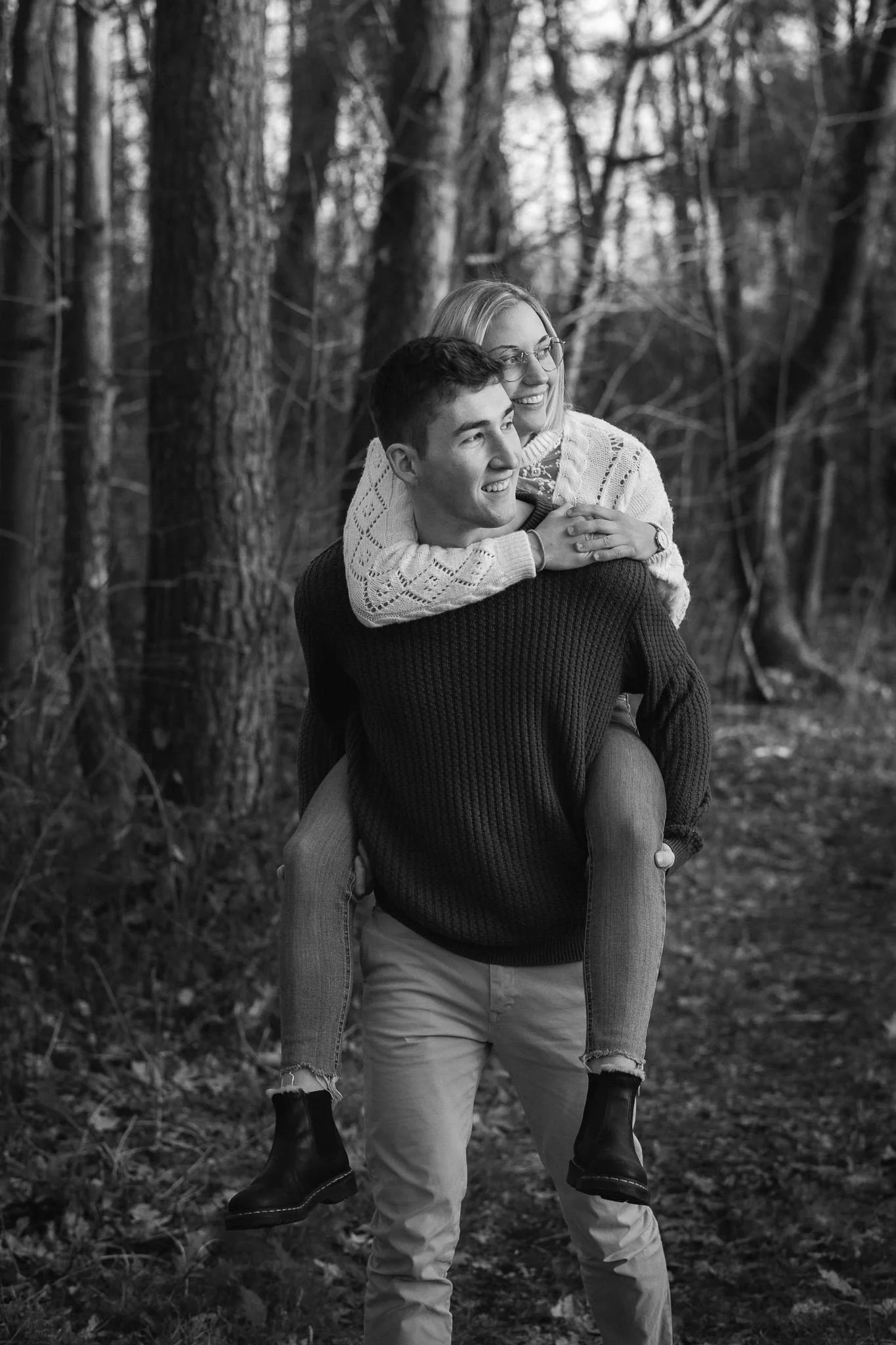 Zwei junge Menschen, ein Mann trägt eine dunkel gestrickte Pullover, darüber eine junge Frau mit Brille und weißem Pullover, sitzen auf den Schultern des Mannes, während sie im Wald spazieren gehen.