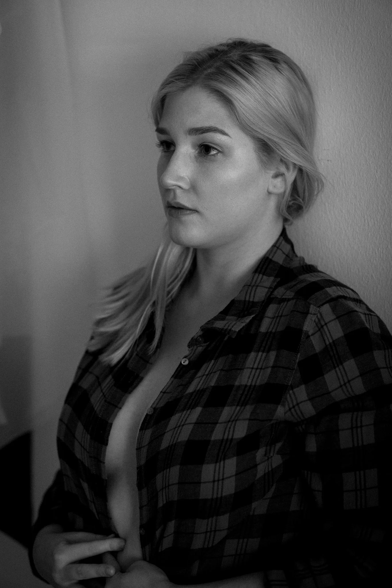 Schwarz-weiß-Fotografie einer jungen Frau, die eine offene, karierte Hemd trägt, seitlich gegen eine Wand lehnend, mit nachdenklichem Ausdruck.
