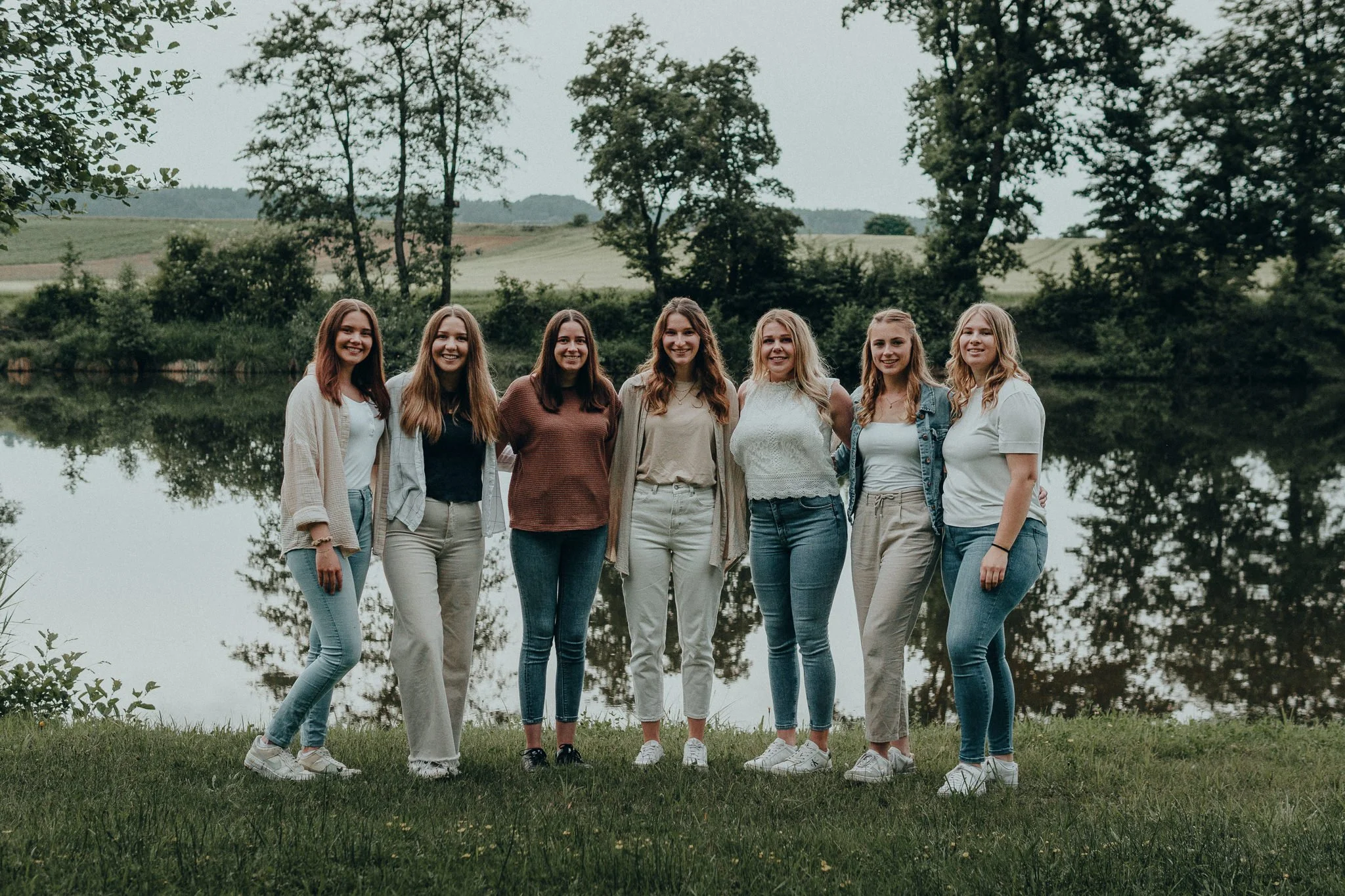 Sieben lachende junge Frauen stehen nebeneinander am Flussufer in einem grünen Park, umgeben von Bäumen und Hügeln im Hintergrund.