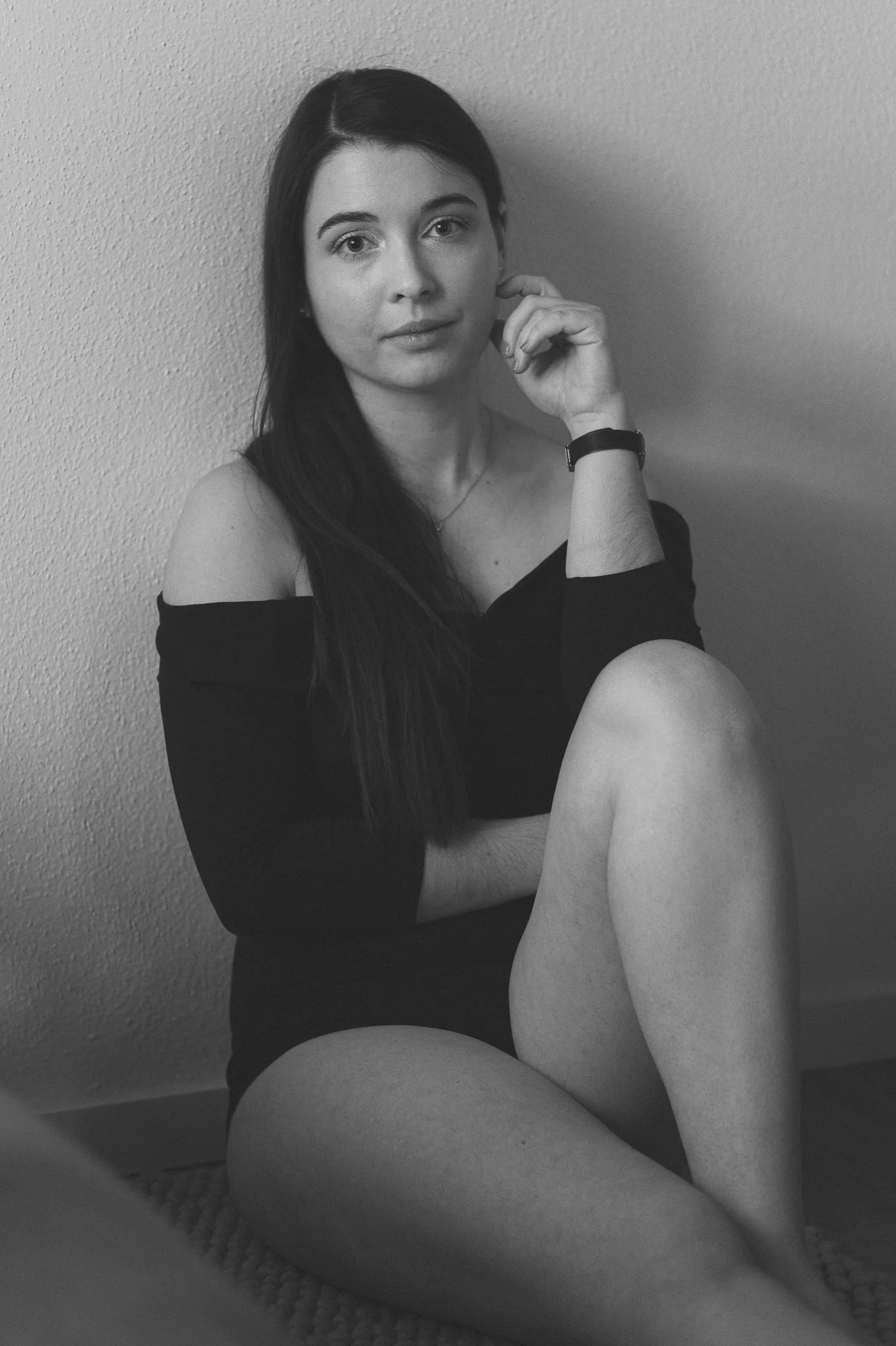 Schwarz-Weiß-Fotografie einer jungen Frau mit langen dunklen Haaren, sitzend am Boden, gekleidet in ein off-shoulder Schwarzkleid, Blick direkt in die Kamera, bei Tag, einfache Umgebung mit glatter Wand im Hintergrund.