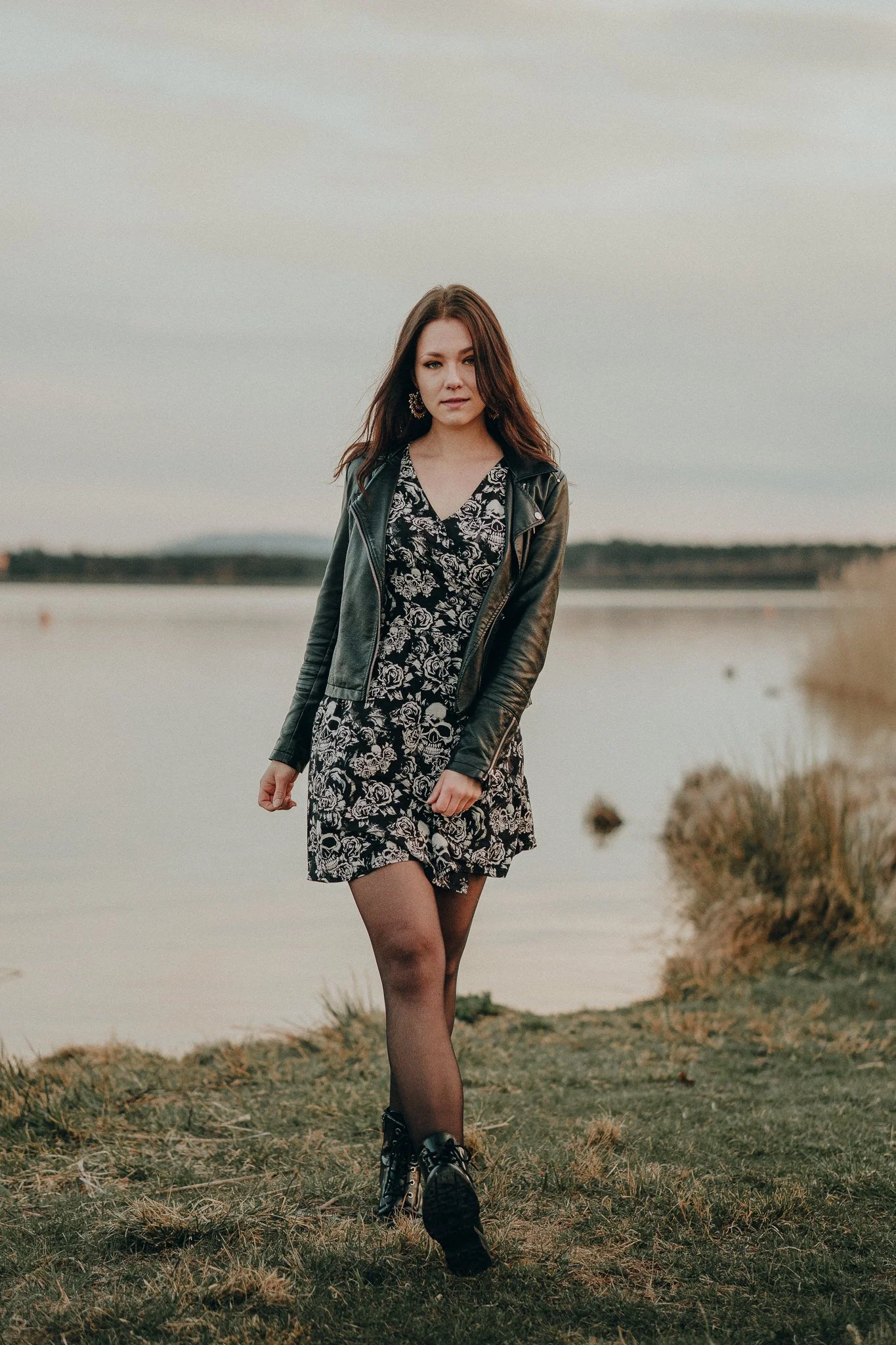 Eine junge Frau läuft an einem See entlang, trägt ein schwarzes Kleid mit weißen Mustern, eine schwarze Lederjacke, schwarze Strümpfe und Stiefel, bei bewölktem Himmel.