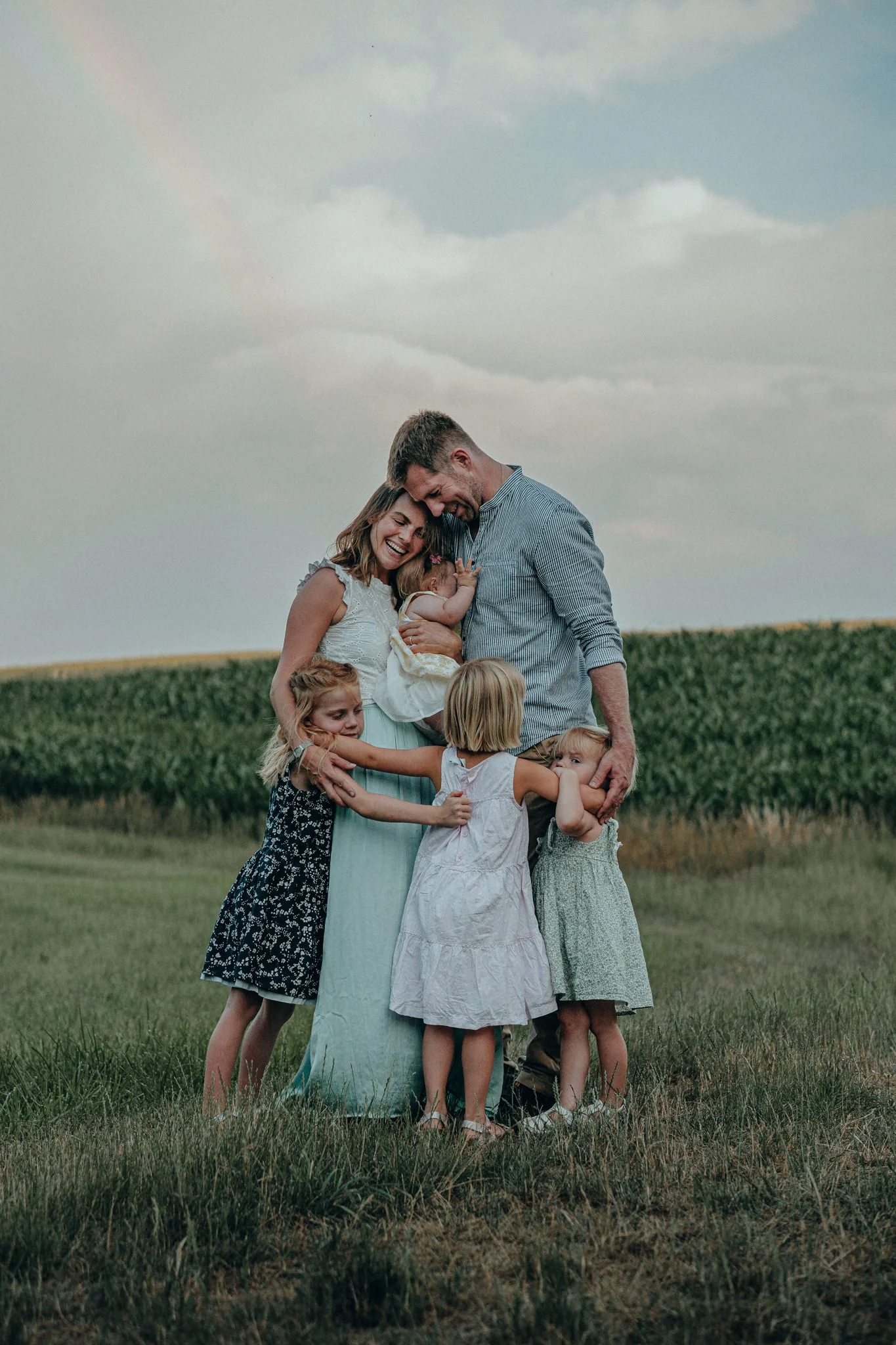 Familie mit zwei erwachsenen Erwachsenen und vier Kindern in einem Feld bei Sonnenuntergang, umarmend und lachend.