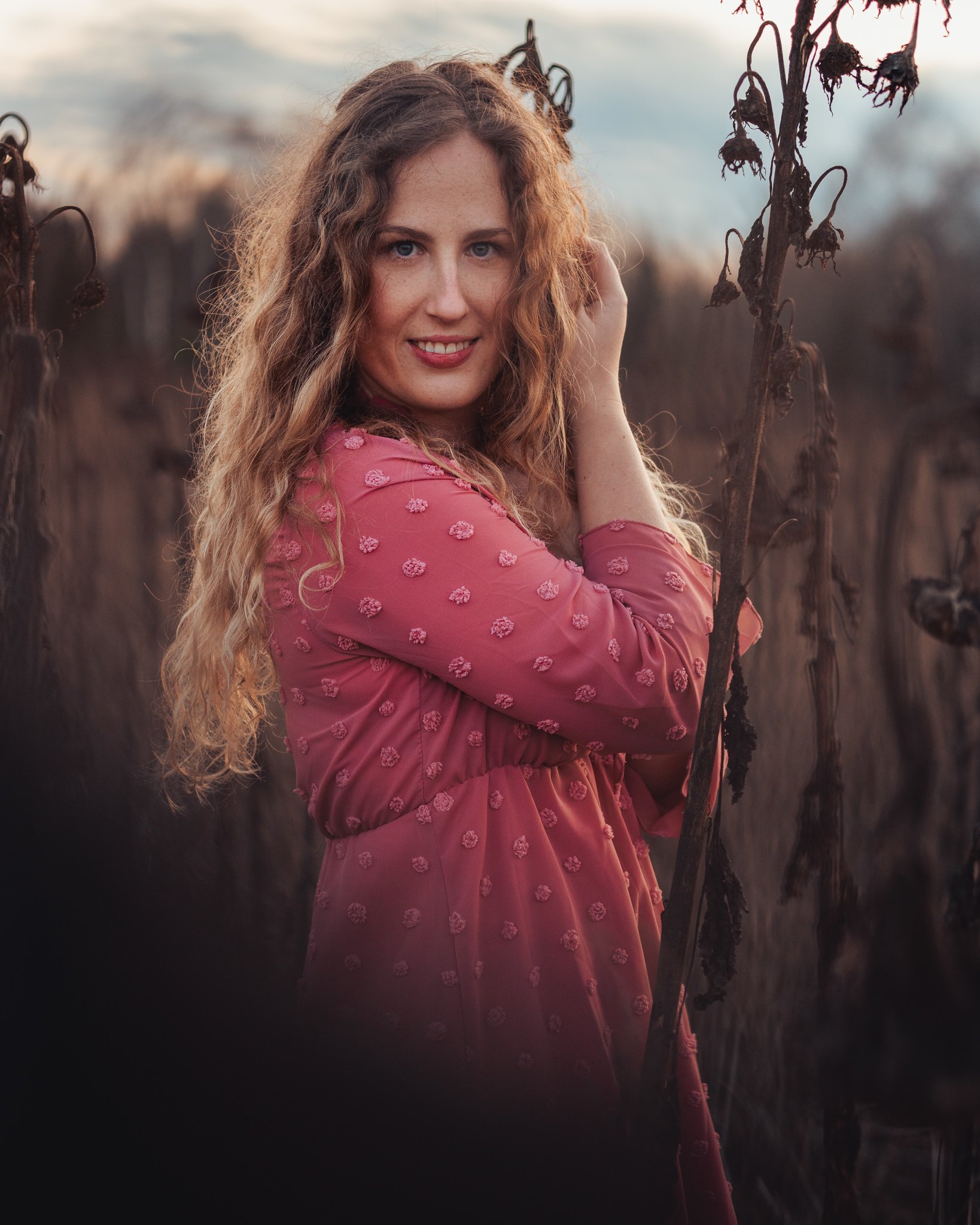 Eine junge Frau mit lockigem, langem Haar in einem pinkfarbenen Kleid mit Muster posiert in einem Feld mit vertrockneten Pflanzen bei Sonnenuntergang.