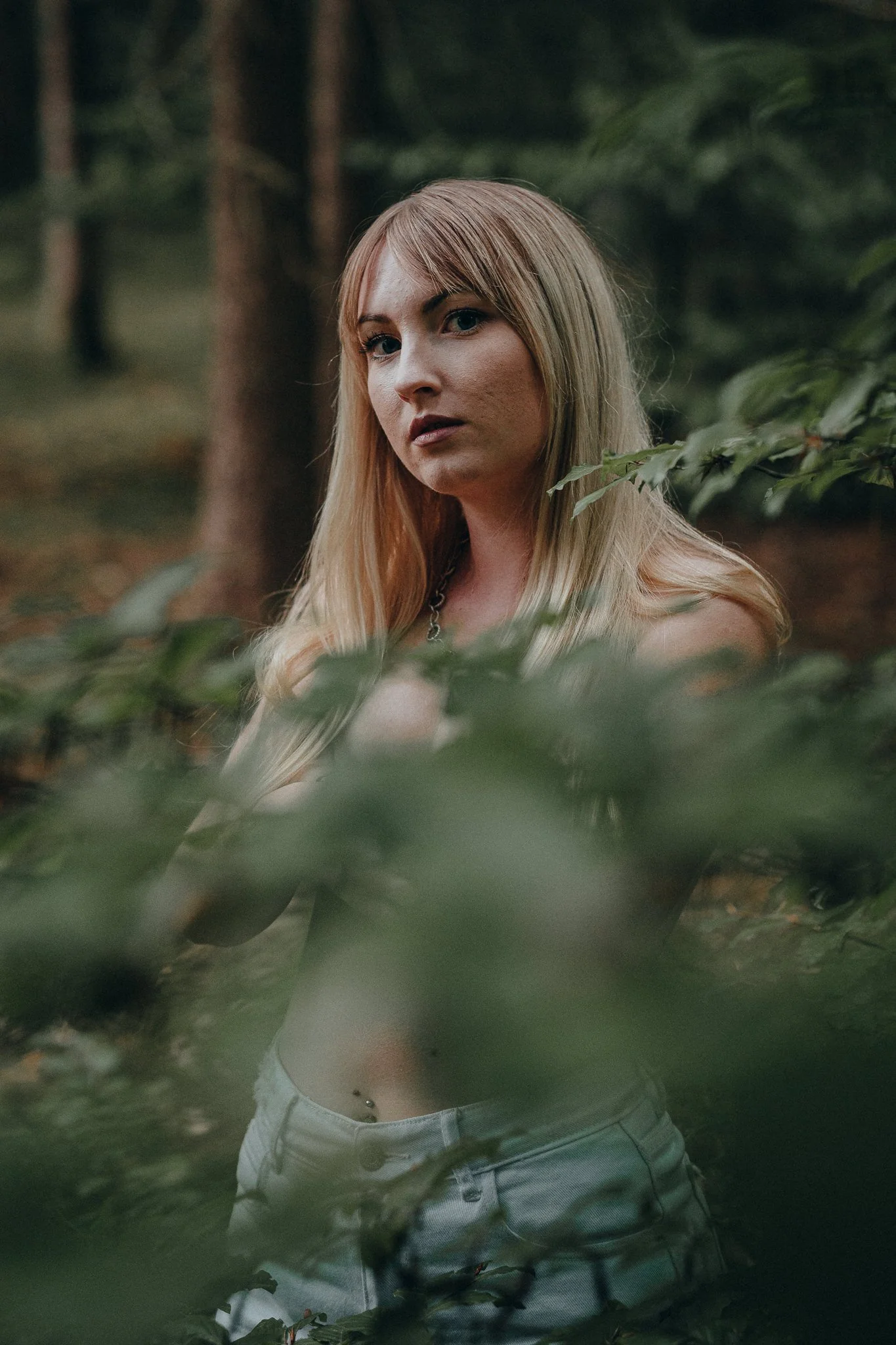 Junge Frau mit blonden Haaren und Piercing im Bauchnabel, die im Wald zwischen Blättern steht.