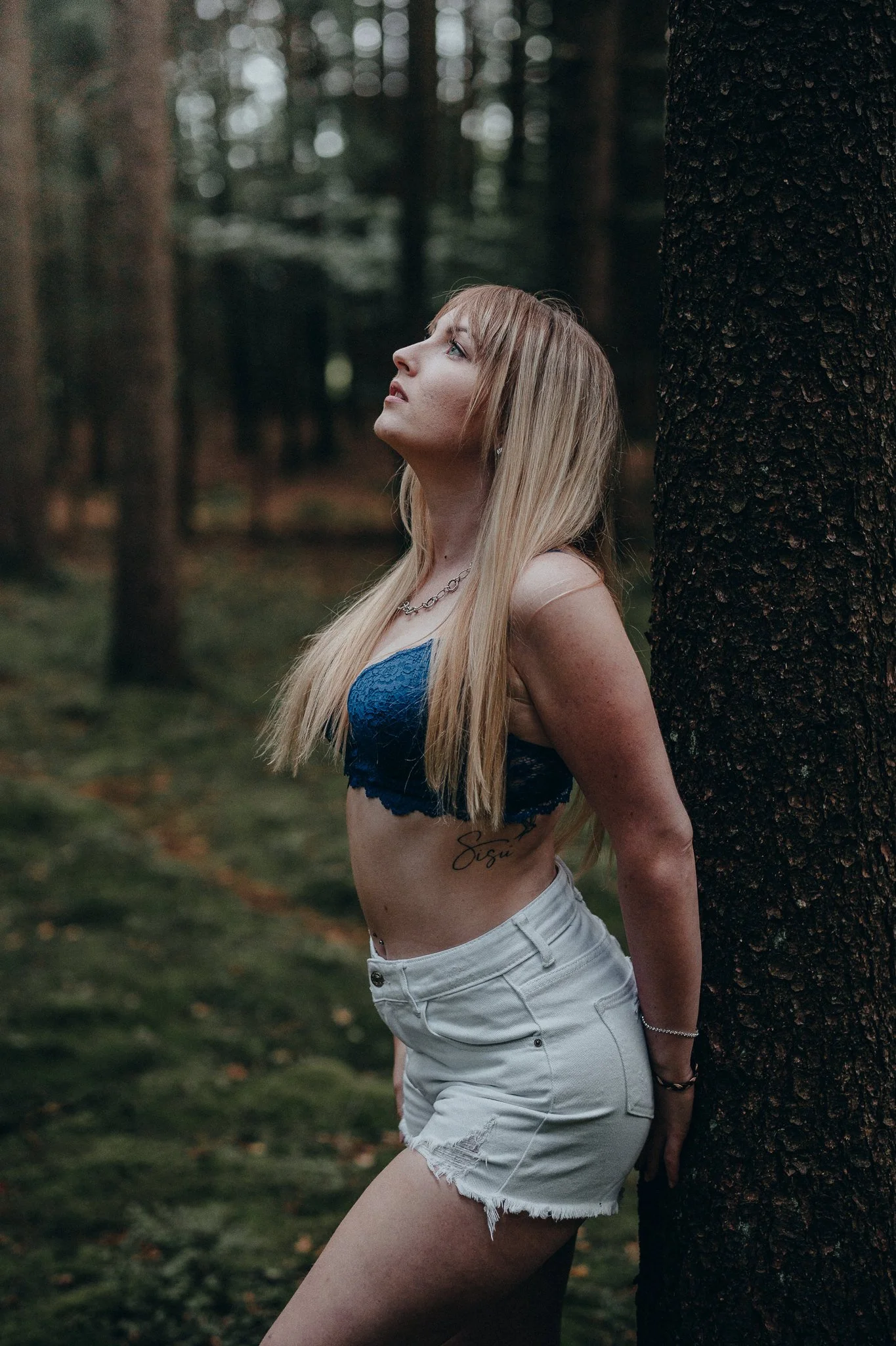 Junge Frau lehnt an Baumstamm im Wald, trägt blauen Top und weiße Shorts, schaut nach oben.