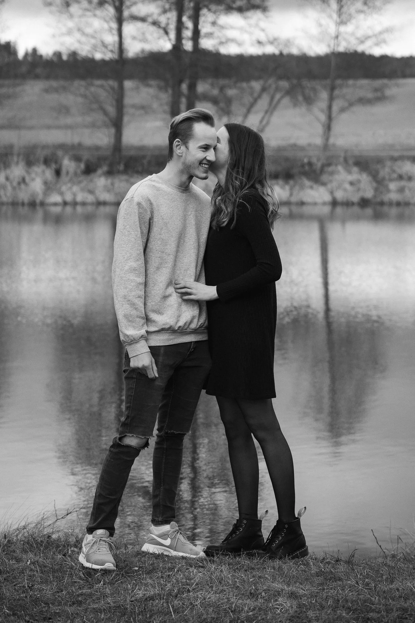 Ein glückliches Paar steht am Ufer eines Sees, der Mann lächelt und die Frau küsst ihn auf die Wange.