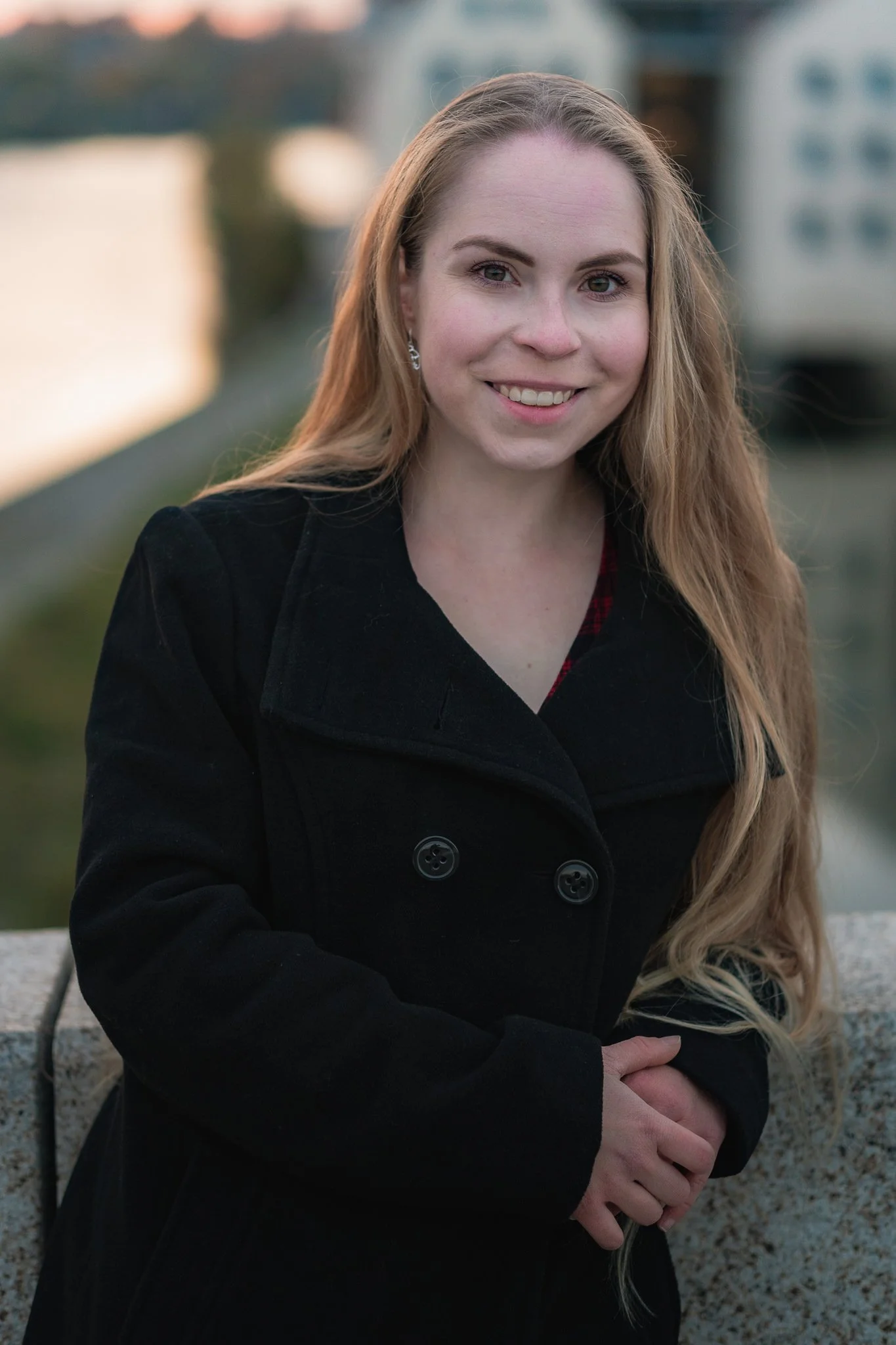 Eine junge Frau mit langen, blonden Haaren, die draußen an einem Abend sitzt, trägt eine schwarze Jacke und lächelt in die Kamera.