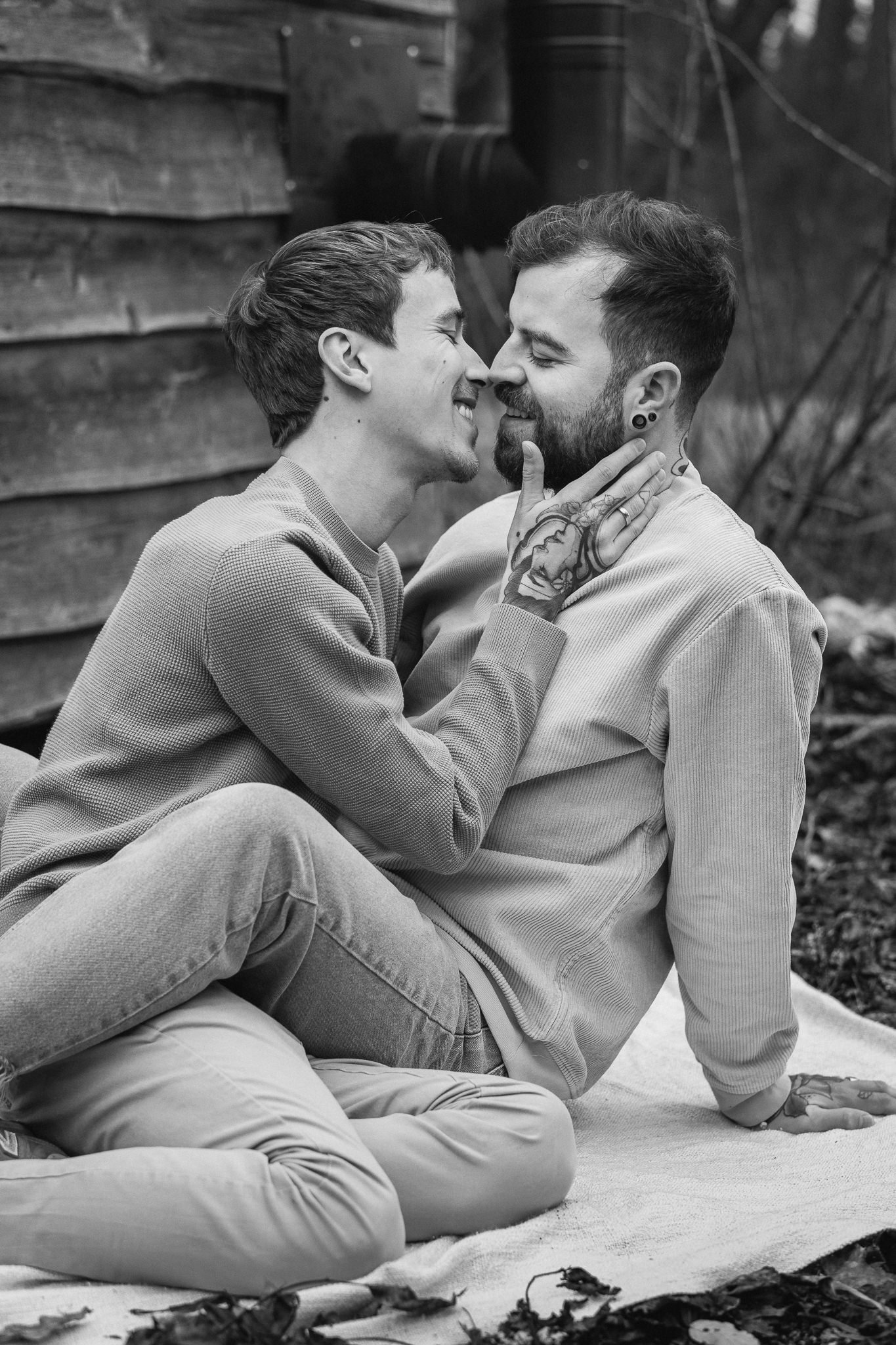 Zwei Männer sitzen sich auf einer Decke im Freien gegenüber, lachen und berühren sich liebevoll an den Gesichtern, während sie sich anlächeln.