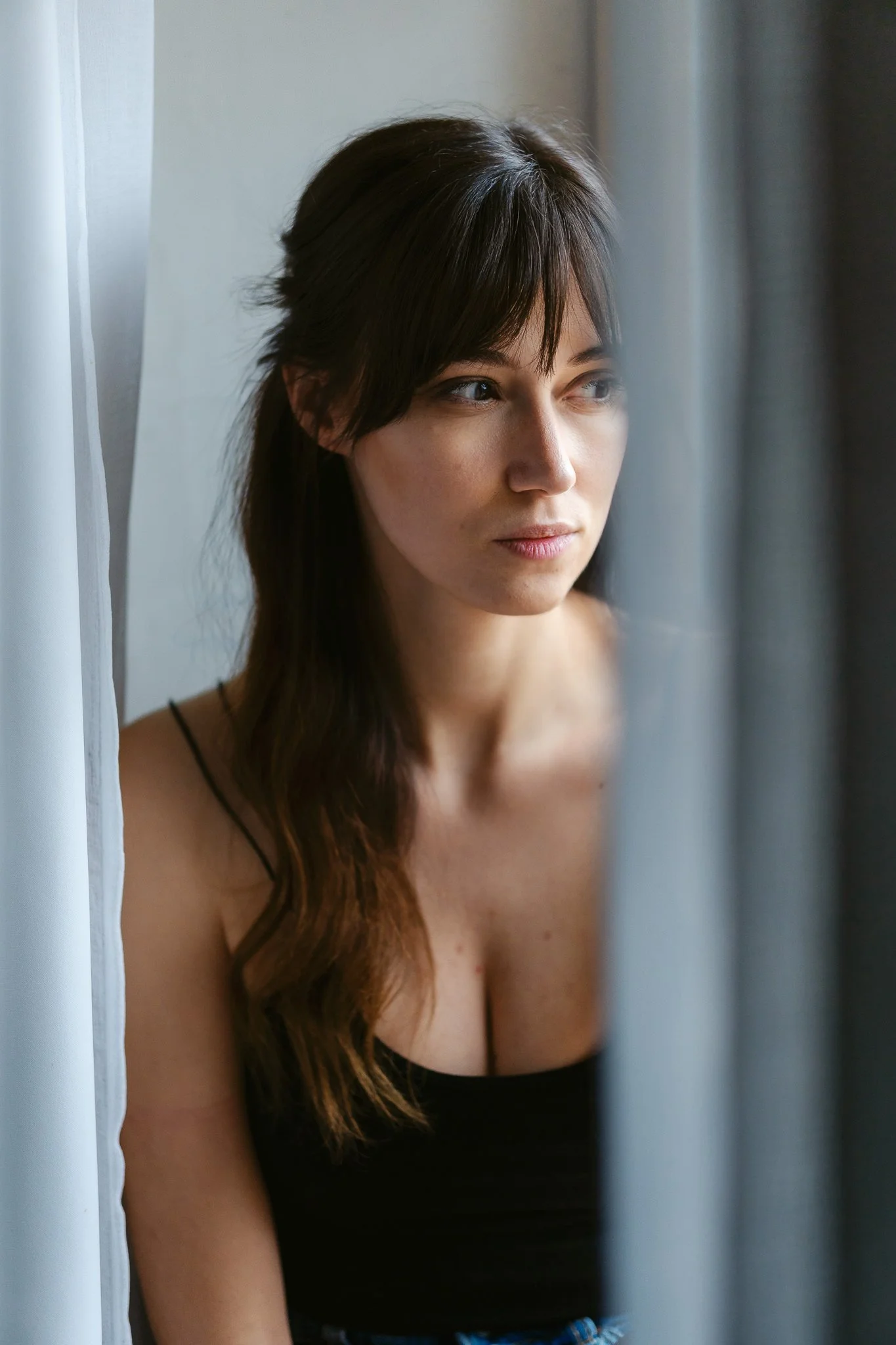 Junge Frau schaut nachdenklich aus einem Fenster, durch einen Vorhang.